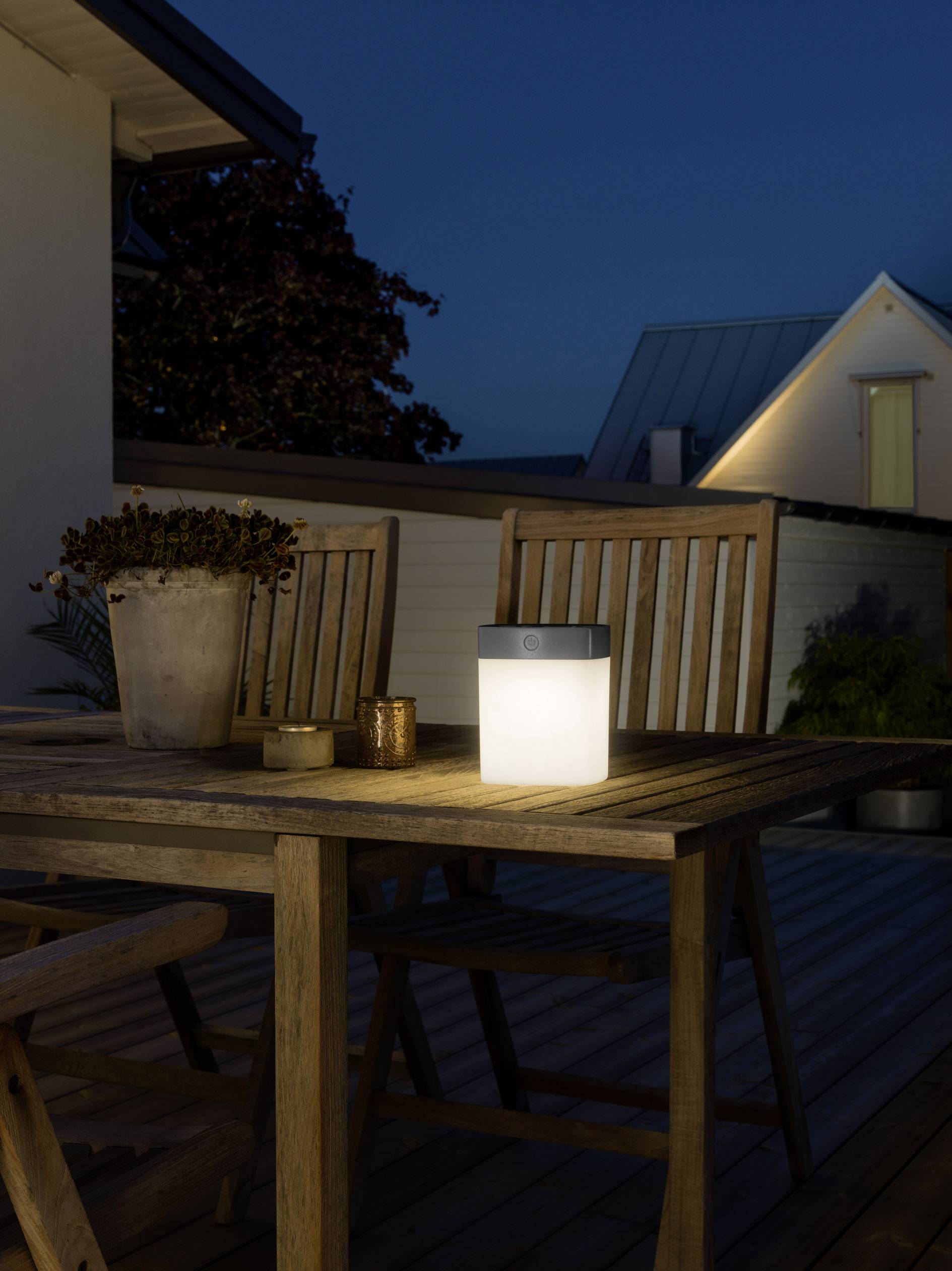 Eine Laterne steht auf einem Holztisch auf einer Terrasse, umgeben von Stühlen. Es ist dunkel, und das Licht der Laterne erhellt die Szene.