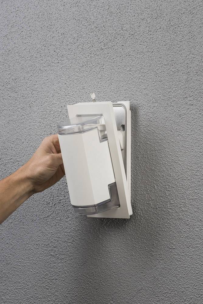 Eine Hand wechselt einen weißen Duftspender, der an einer grauen Wand montiert ist.