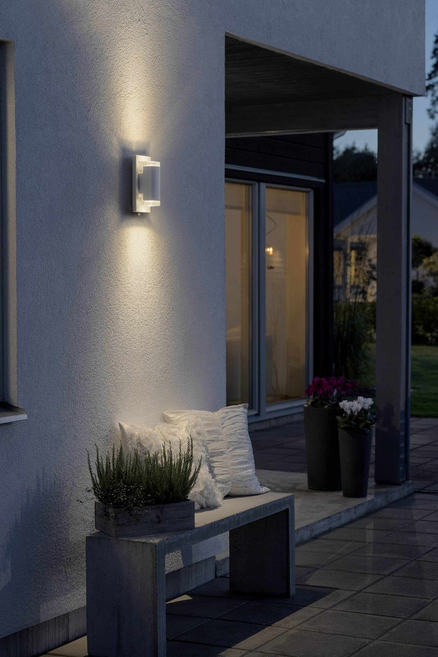 Wandleuchte an einer Hauswand, beleuchtet abends eine Terrasse mit Sitzbank und Pflanzen. Im Hintergrund sind Fenster und ein Garten sichtbar.
