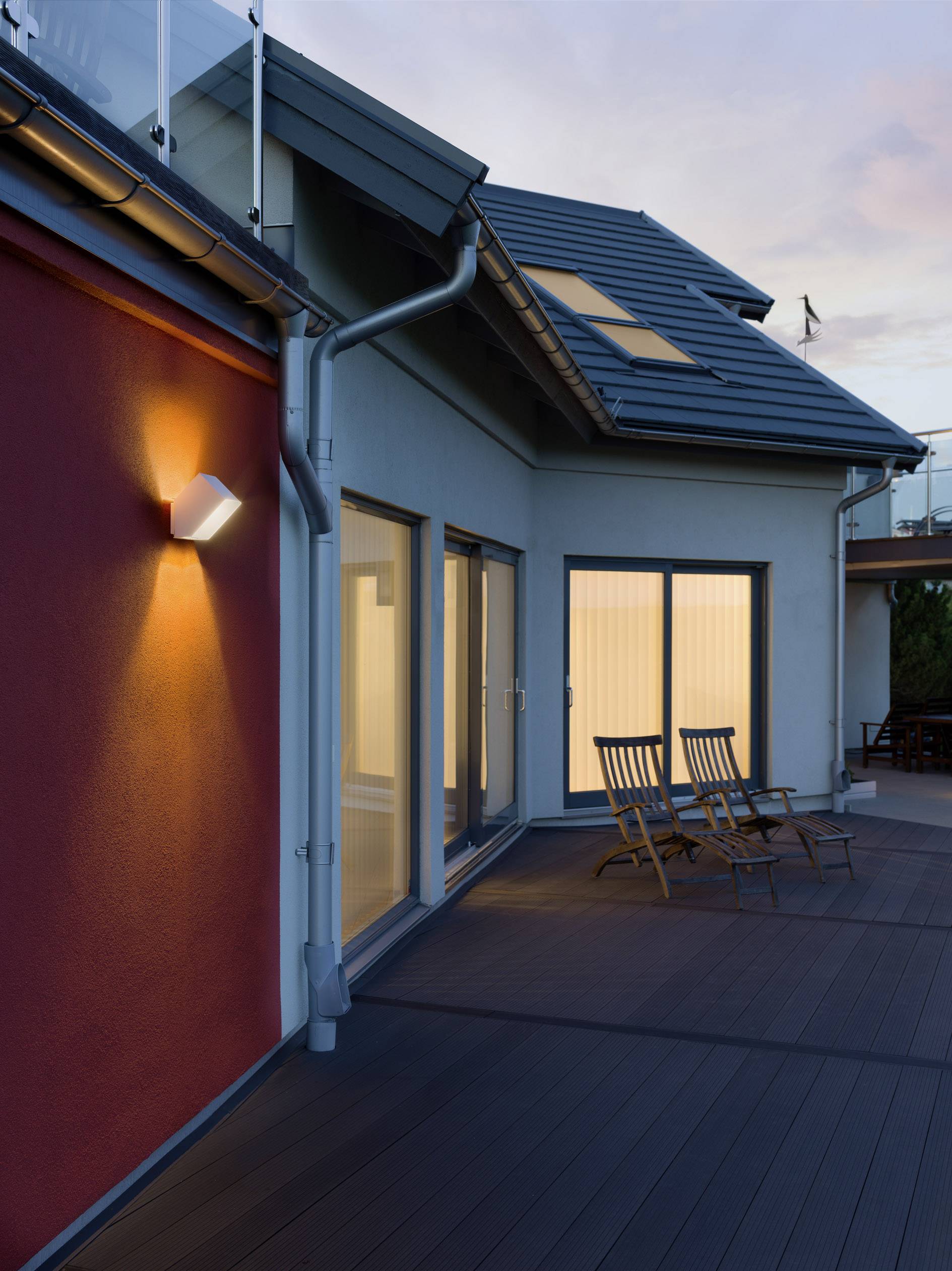 Abendaufnahme einer modernen Terrasse mit zwei Liegestühlen vor einem Haus. Warmes Licht strahlt von einer Lampe an der roten Wand.