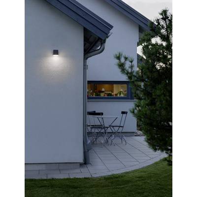 Ein modernes Haus mit eingeschalteter Außenbeleuchtung. Auf der Terrasse stehen ein Tisch und zwei Stühle. Ein Fenster zeigt Blumen im Inneren.