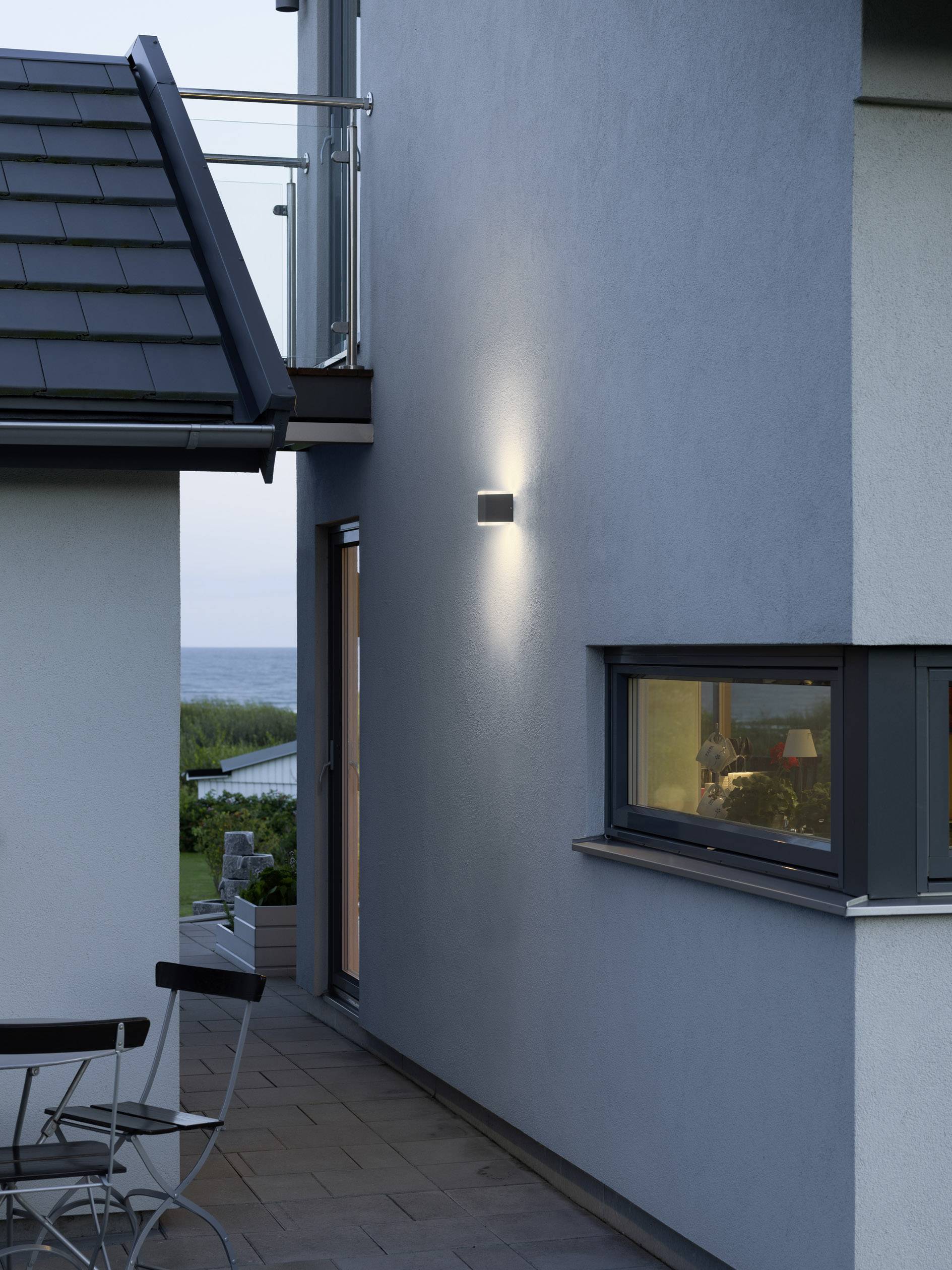 Moderner Außenbereich eines Hauses in der Dämmerung, mit beleuchteter Außenwand und Aussicht auf das Meer im Hintergrund.
