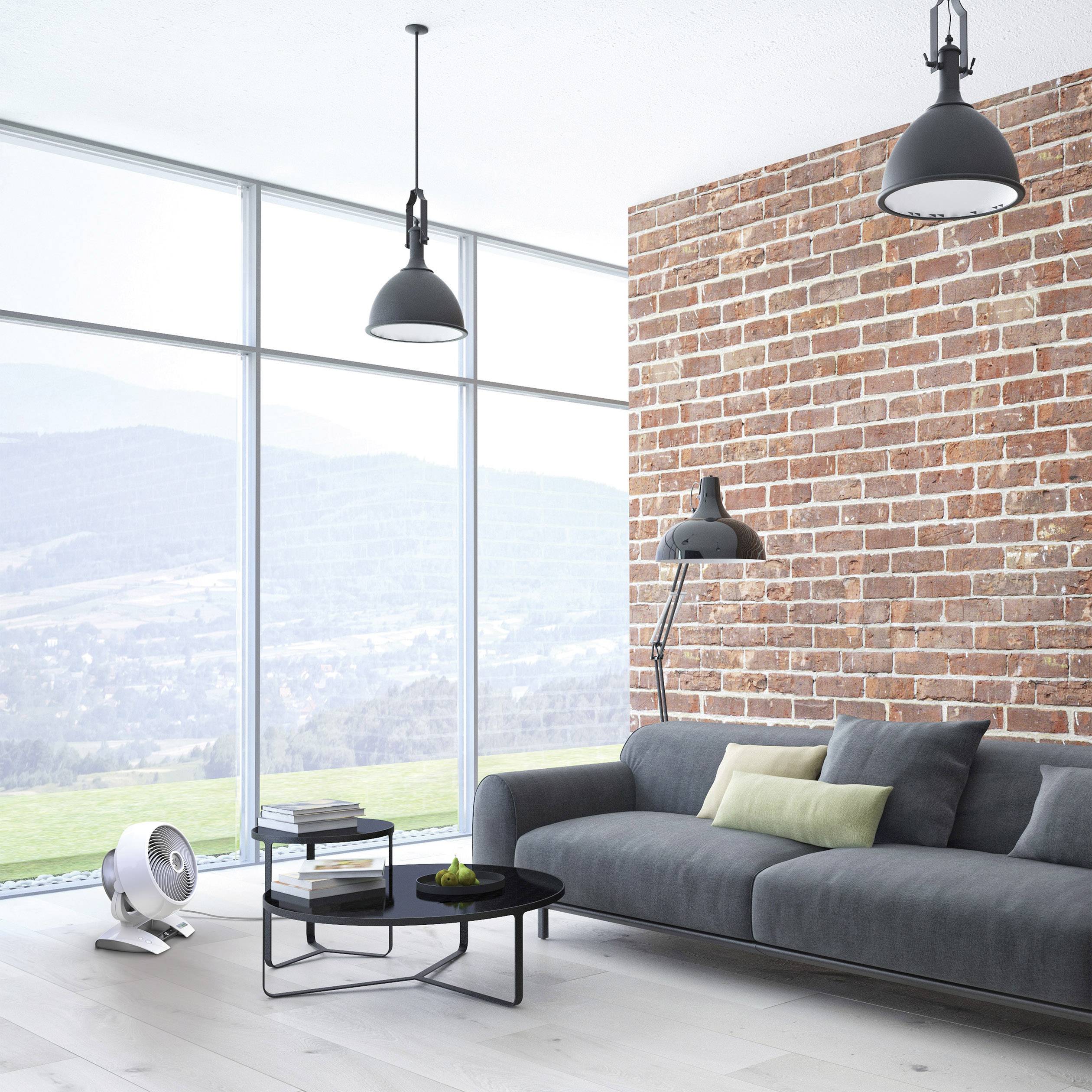 Wohnzimmer mit großer Fensterfront, grauem Sofa vor einer Ziegelwand, zwei schwarzen Deckenlampen, einem kleinen Tisch und einem Ventilator.
