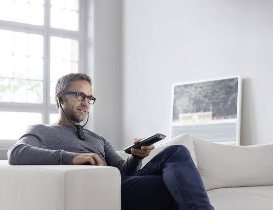 Ein Mann hört mit Kopfhörern Musik und hält eine Fernbedienung, während er entspannt auf einem weißen Sofa sitzt.