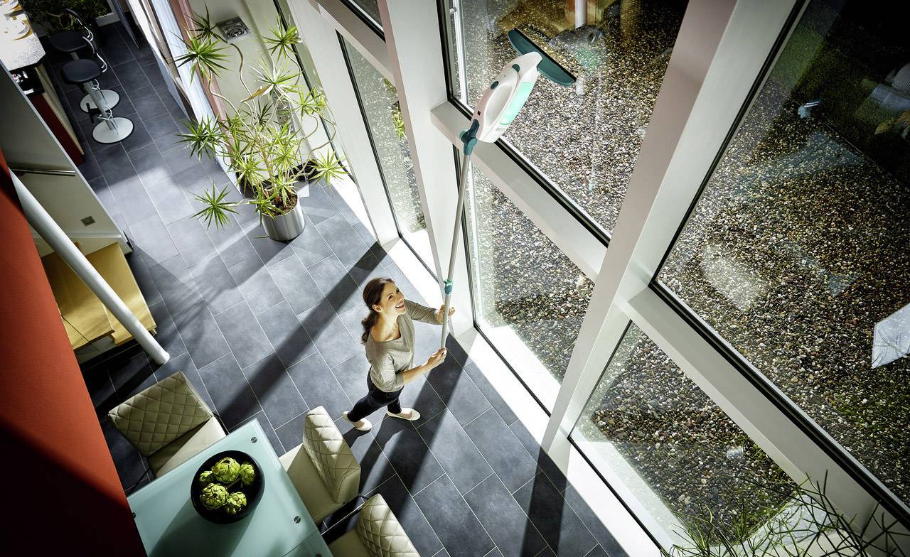 Frau reinigt mit einem langen Gerät die großen Fenster in einem modernen Raum. Pflanzen und moderne Einrichtung sind zu sehen.