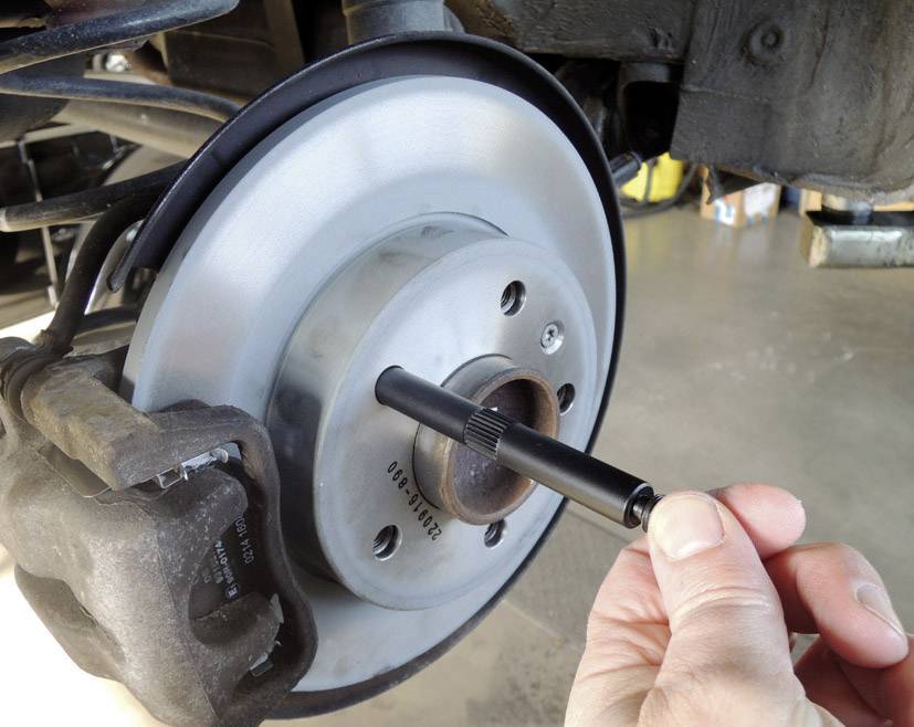 Hand hält ein Diagnosetool an die Bremsscheibe eines Autos, um die Dicke zu messen. Schraubenschlüssel und andere Teile sind sichtbar.
