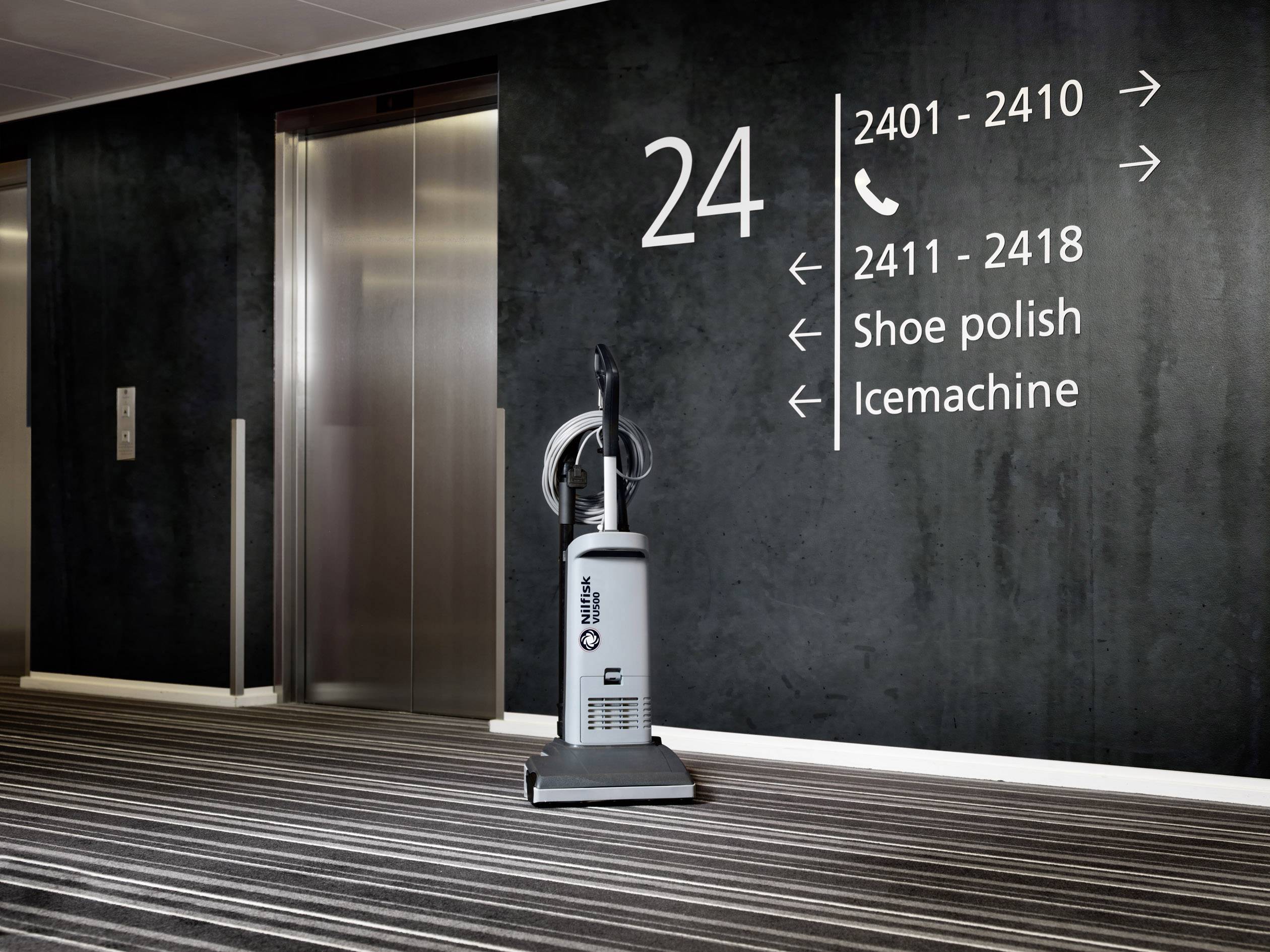 Ein grauer Staubsauger steht auf einem gestreiften Teppich vor einer schwarzen Wand mit weißen Zimmernummern und Hinweisschildern.