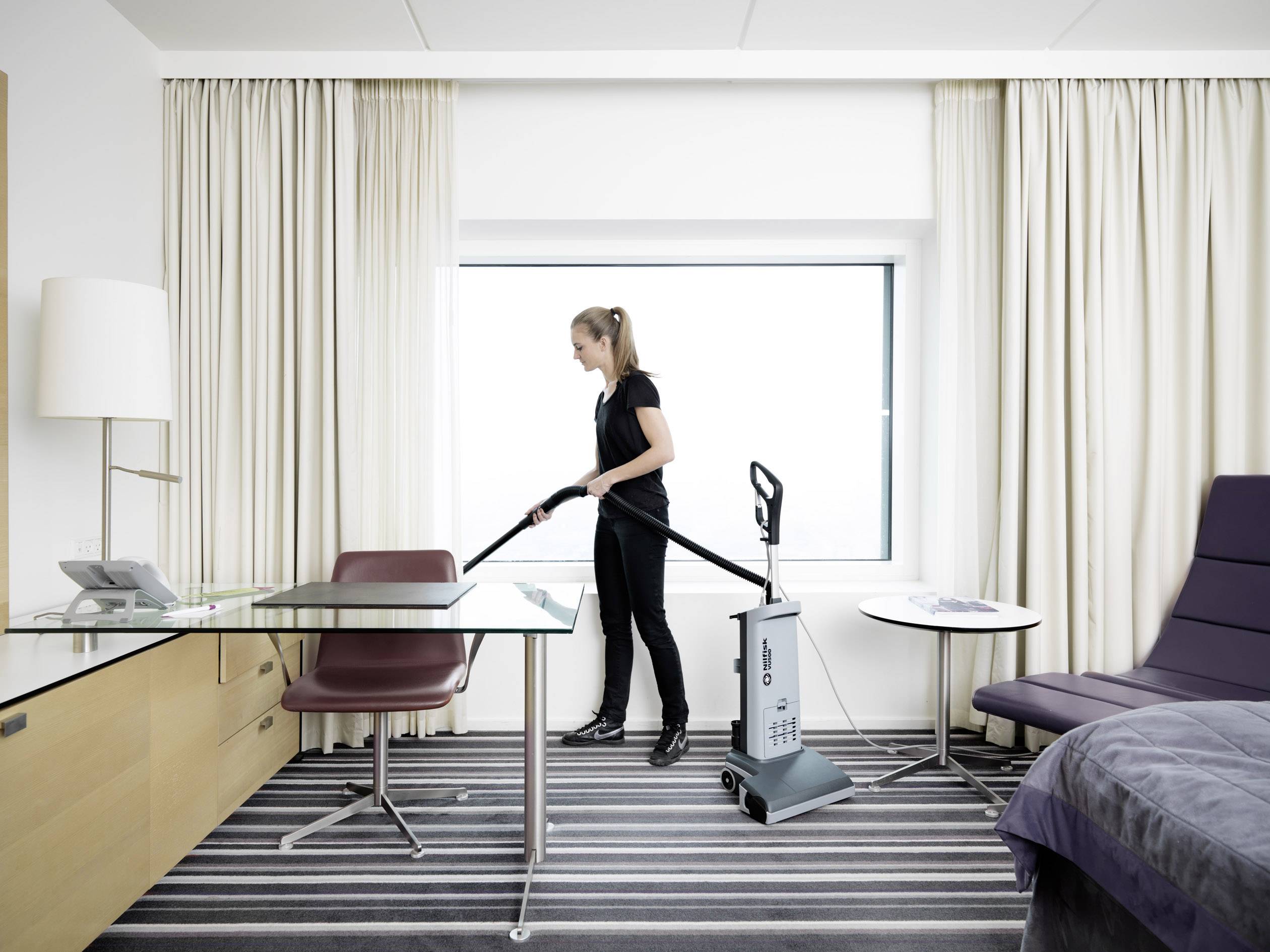 Eine Person reinigt mit einem Staubsauger ein modernes Hotelzimmer, das einen Glastisch, einen Stuhl und einen Sessel enthält.