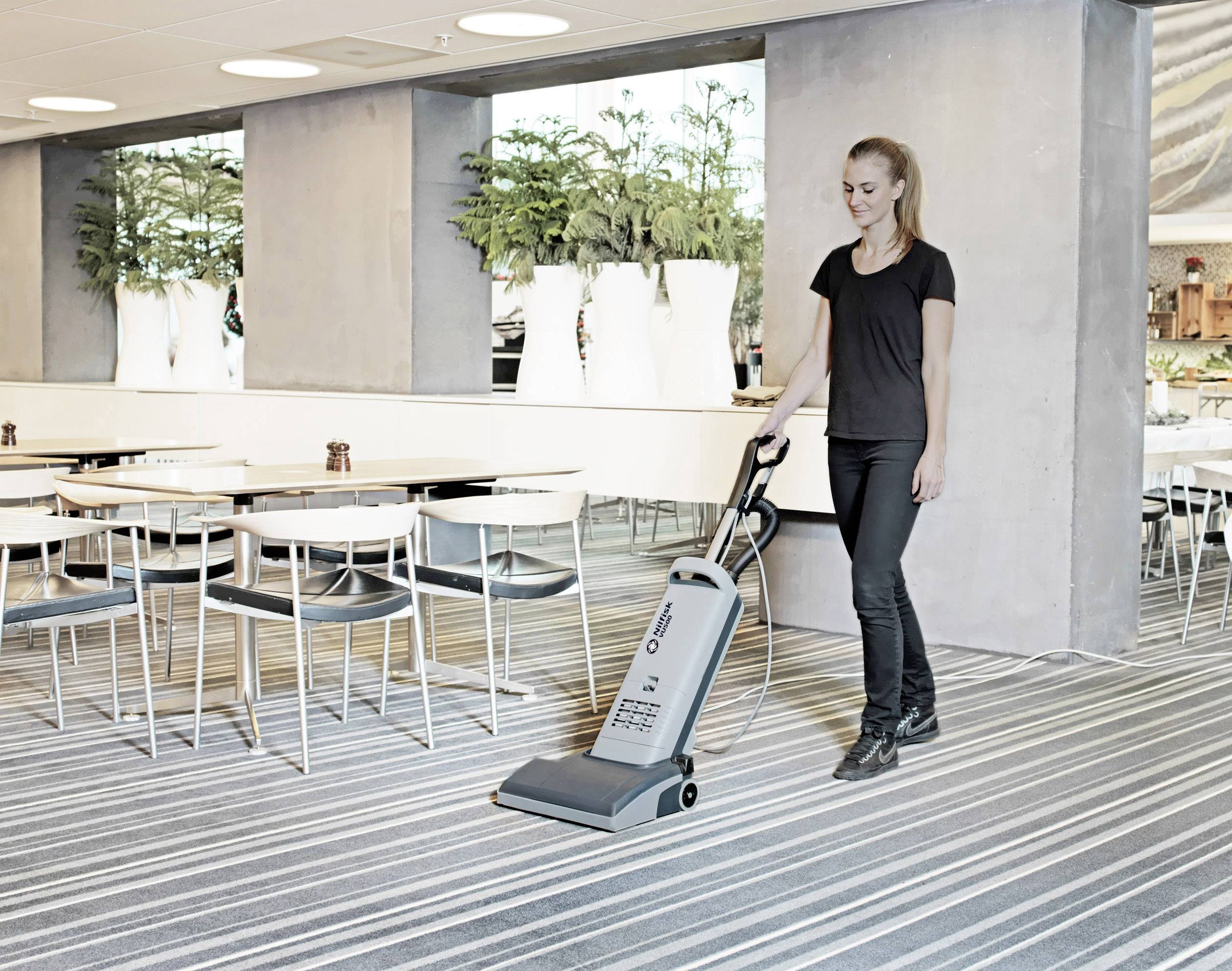 Eine Person reinigt den Teppichboden in einem modernen Speisesaal mit einem Staubsauger. Im Hintergrund stehen Tische und Stühle.