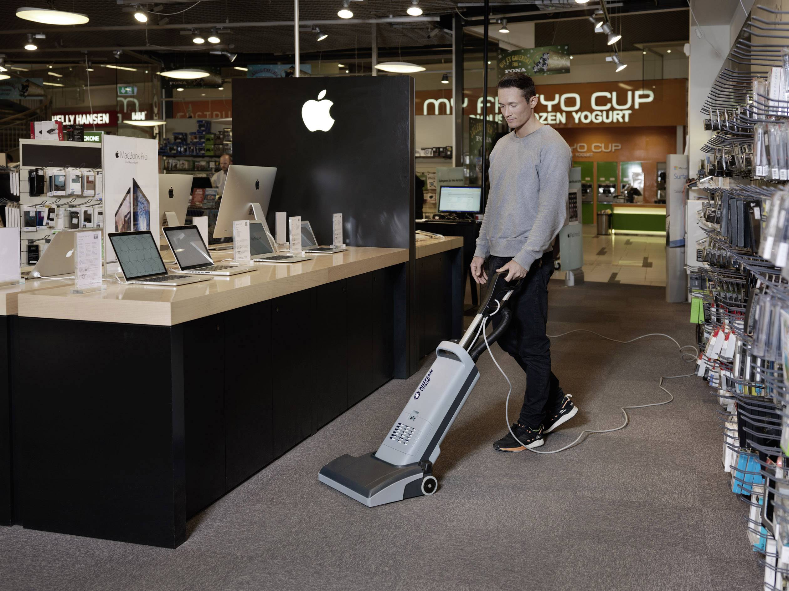 Eine Person staubsaugt den Boden in einem Elektronikgeschäft. Auf einem Tisch stehen verschiedene Laptops und Tablets. Im Hintergrund ist ein Apple-Logo sichtbar.