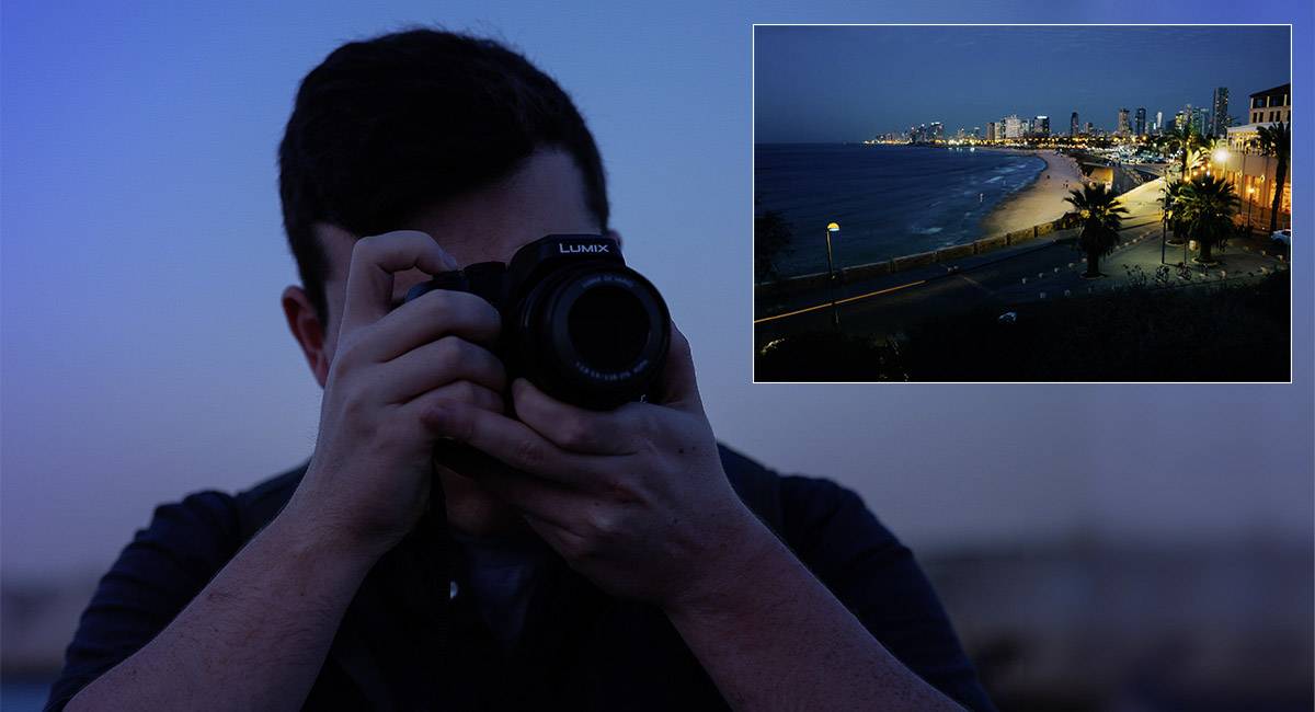 Eine Person fotografiert eine nächtliche Strandpromenade einer Stadt. Im Bild sind Gebäude mit Lichtern und ein beleuchteter Strandweg zu sehen.