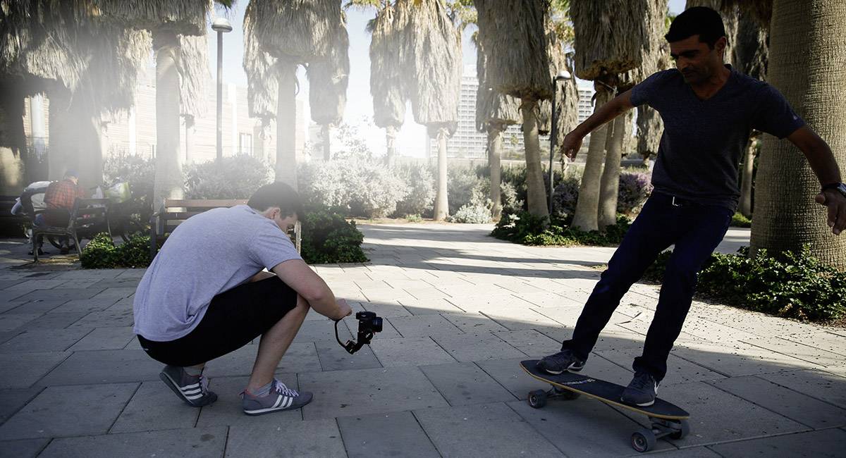 Person auf Skateboard fährt auf Gehweg in einem Park, während eine andere Person daneben in die Hocke geht und mit einer Kamera filmt.