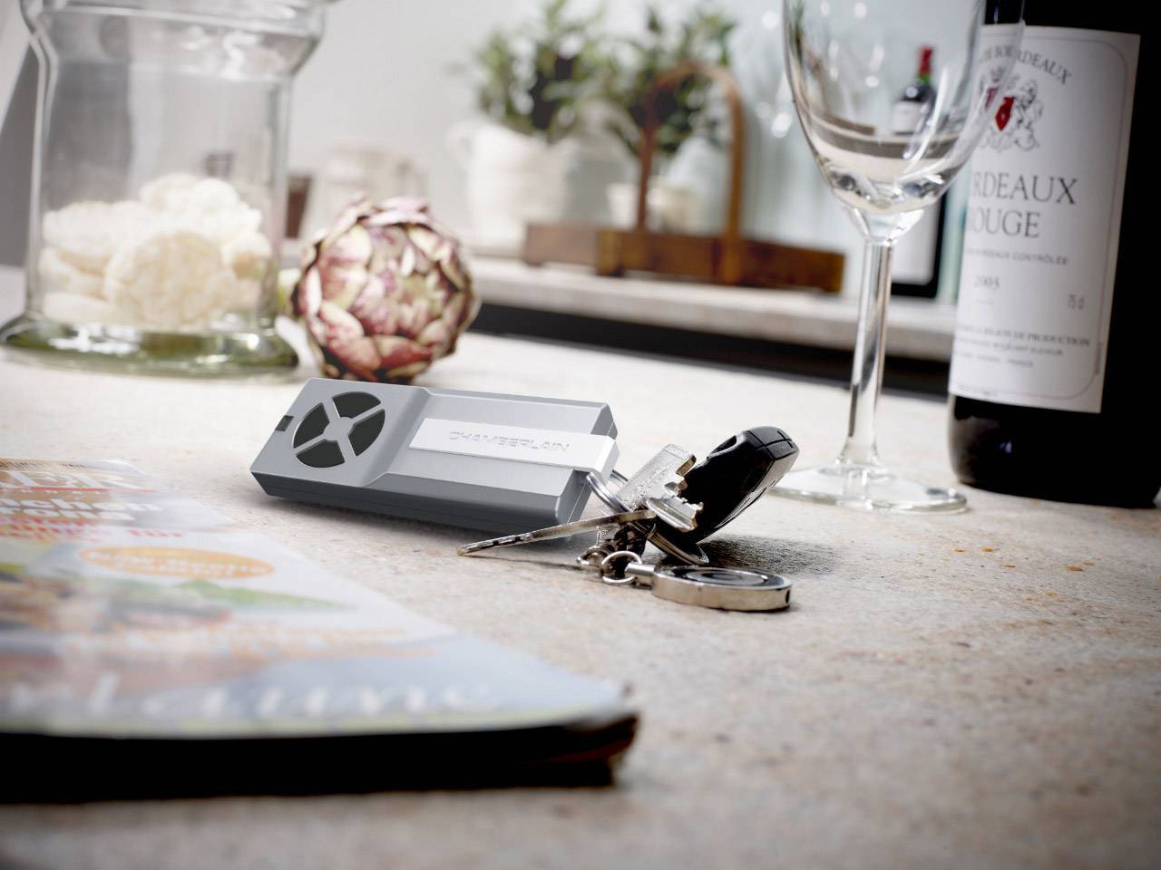 Weinflasche Bordeaux Rouge, Weinglas und Schlüssel auf einem Tisch. Im Hintergrund ein Artischocken-Dekor und Zeitschrift.