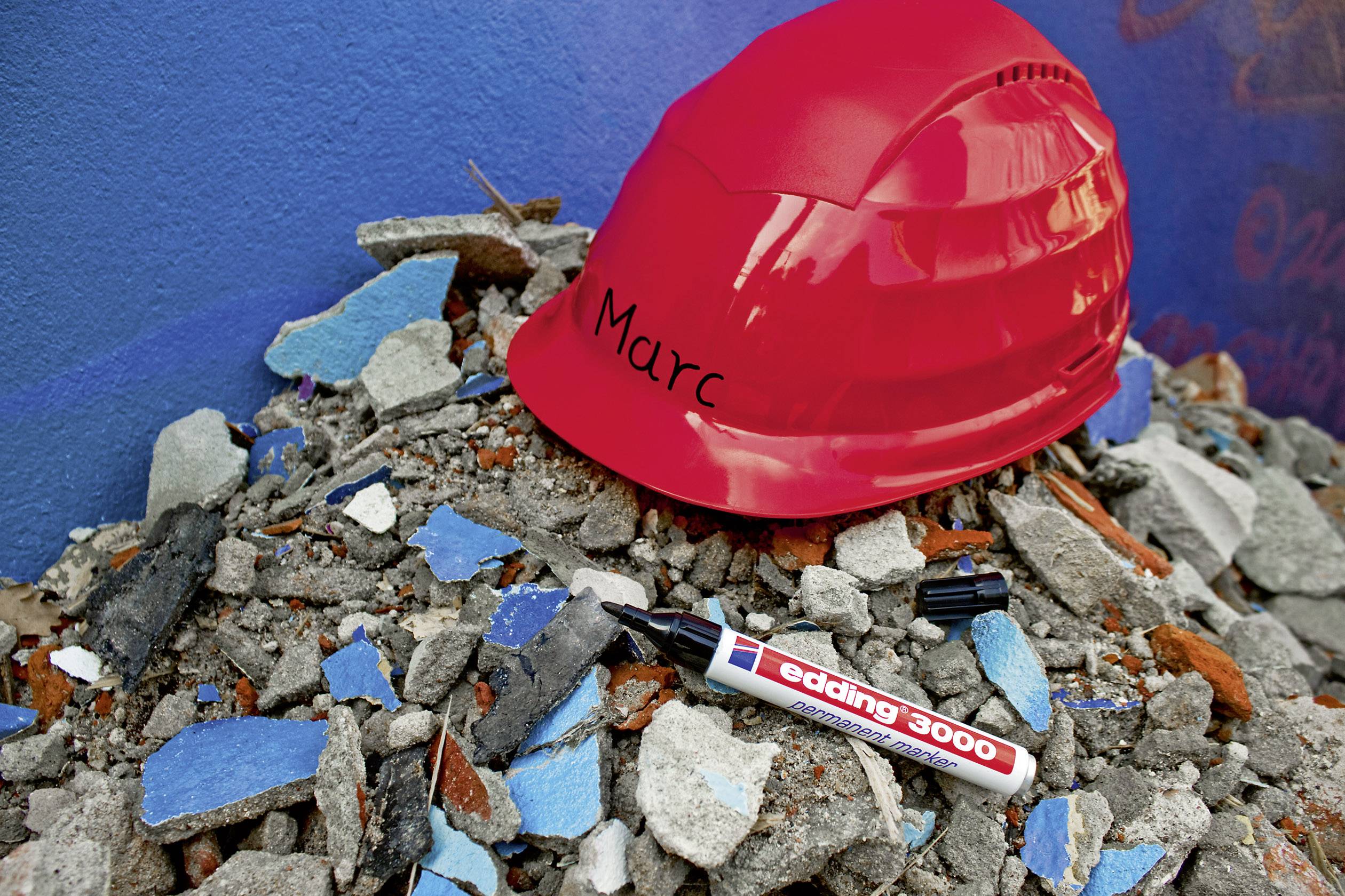 'Roter Bauhelm mit Name Marc, liegt auf einem Trümmerhaufen. Daneben ein Marker, Marke edding 3000. Hintergrund: blaue Wand.'