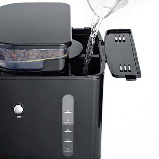 Eine Kaffeemaschine mit Wasserbehälter wird von einer Kanne Wasser befüllt. Oben befindet sich ein Fach mit Kaffeebohnen.