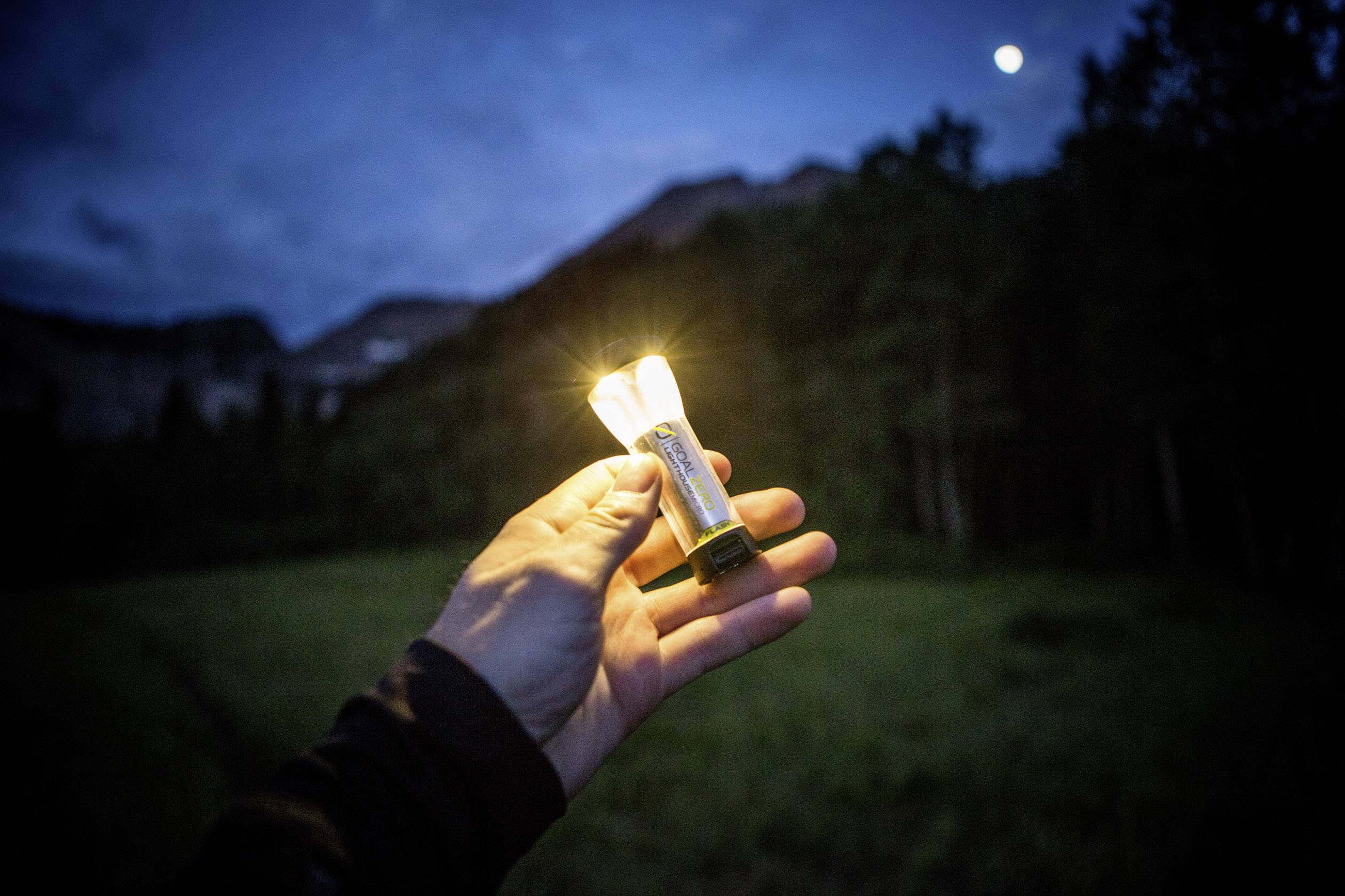 Eine Hand hält eine leuchtende Glühbirne in einer natürlichen Umgebung bei Dämmerung. Im Hintergrund sind unscharfe Bäume und ein Hügel sichtbar.