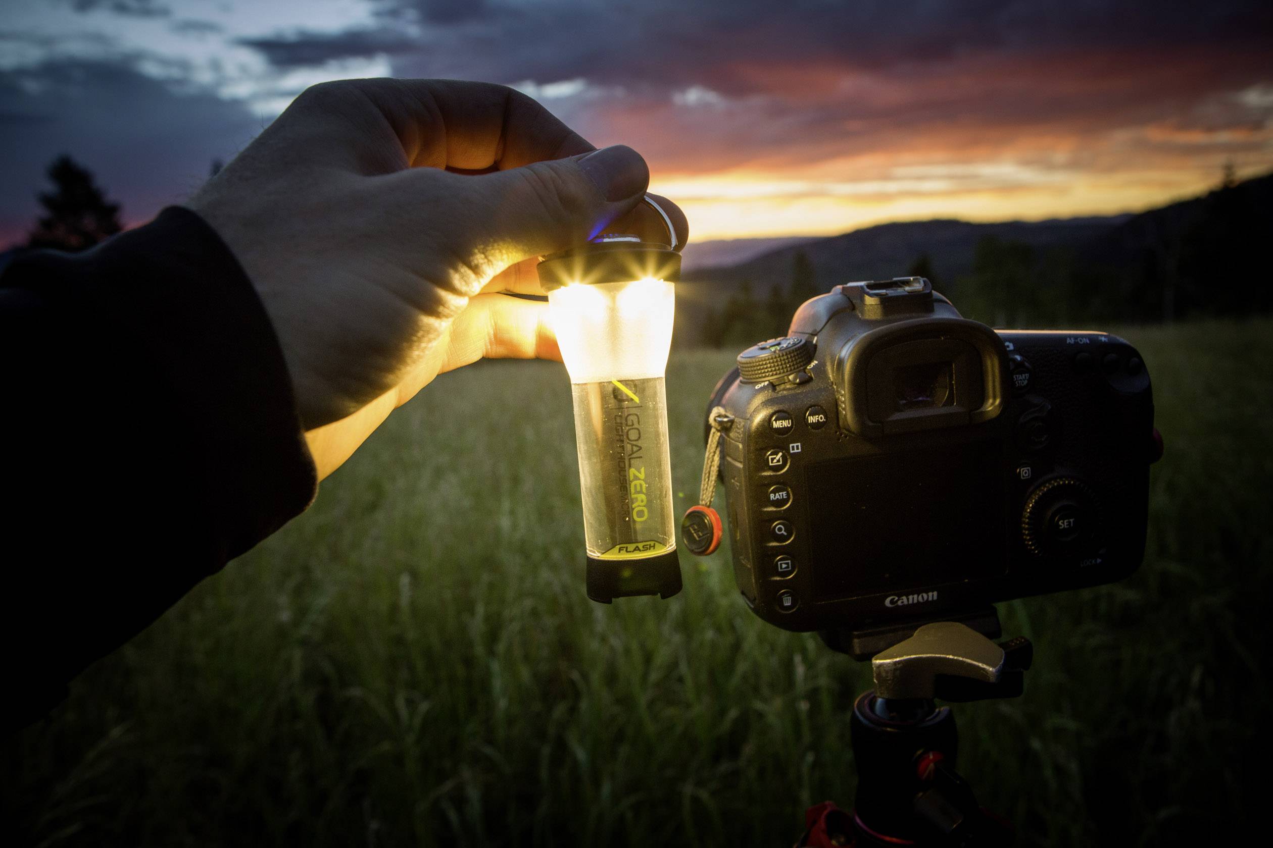 Eine Hand hält eine leuchtende Lampe neben einer Kamera auf einem Stativ, Hintergrund: Sonnenuntergang über einem Feld.