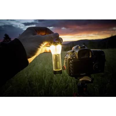 Eine Hand hält eine leuchtende Lampe neben einer Kamera auf einem Stativ, Hintergrund: Sonnenuntergang über einem Feld.