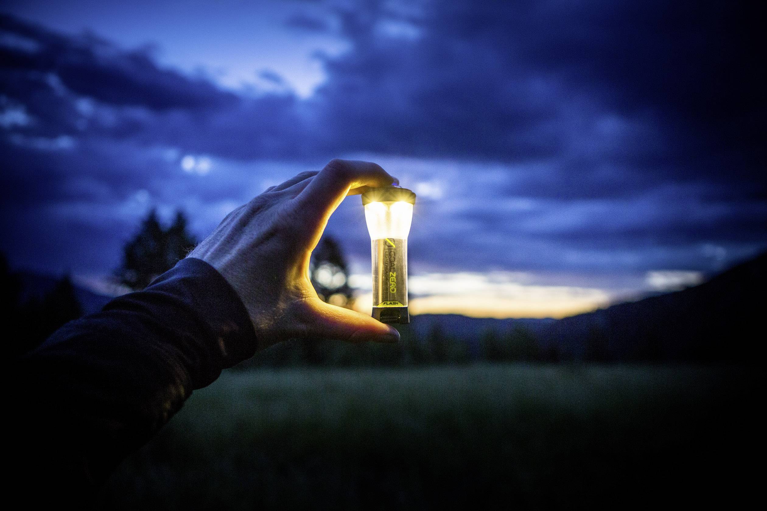 Eine Hand hält eine kleine Taschenlampe vor einem dunklen, wolkigen Himmel in der Dämmerung. Die Lampe leuchtet hell in der Dunkelheit.