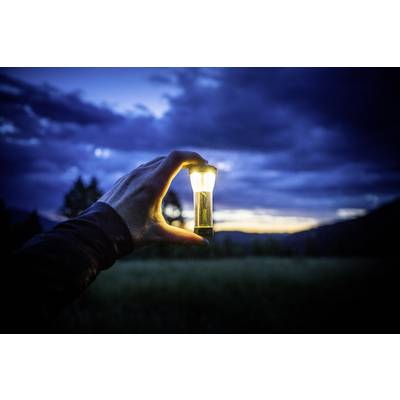 Eine Hand hält eine kleine Taschenlampe vor einem dunklen, wolkigen Himmel in der Dämmerung. Die Lampe leuchtet hell in der Dunkelheit.