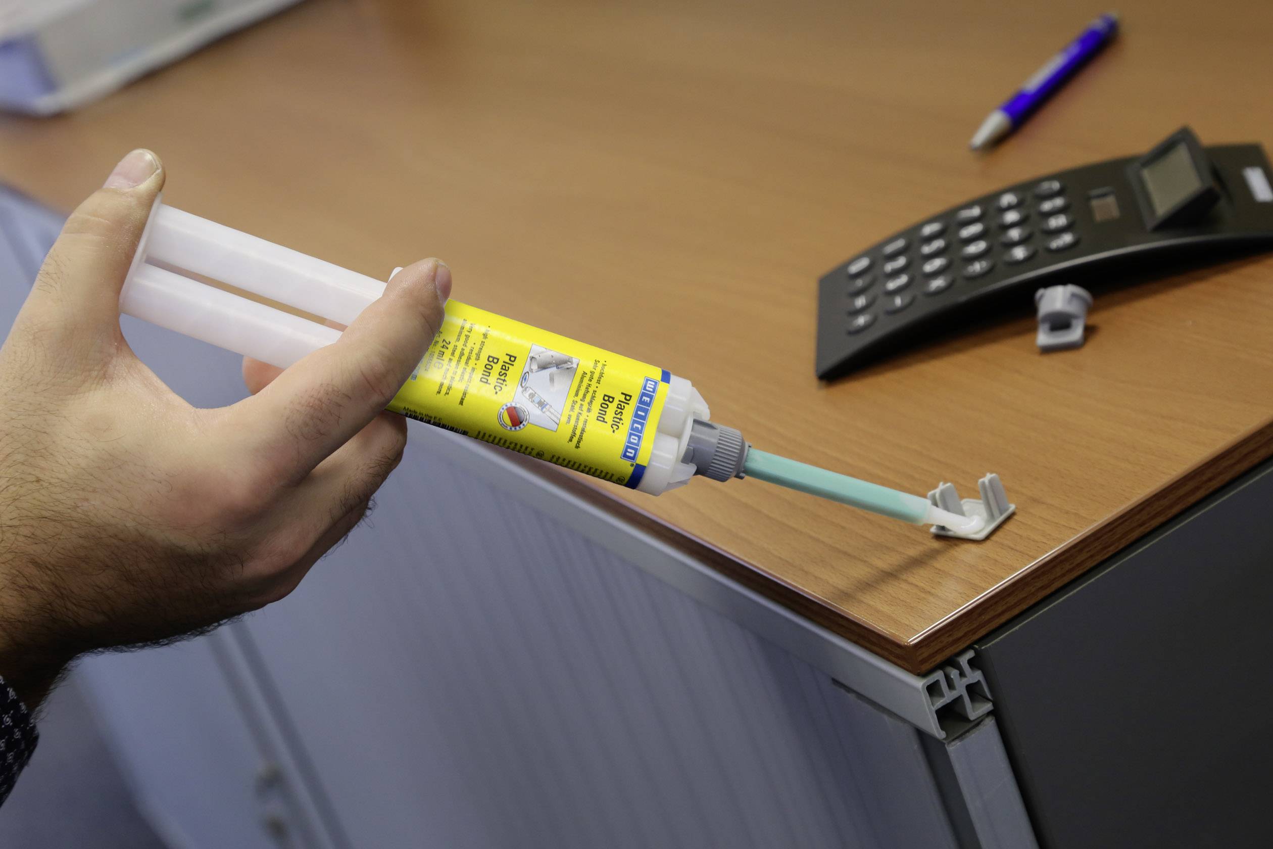 Eine Person trägt Klebstoff mit einer Doppelkartusche auf ein Metallelement an einer Tischkante auf. Auf dem Tisch befindet sich ein Taschenrechner.