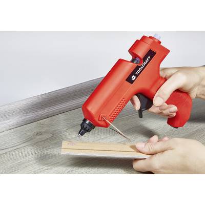 Eine Person verwendet eine rote Heißklebepistole, um ein Holzstück auf einer Tischoberfläche zu kleben.