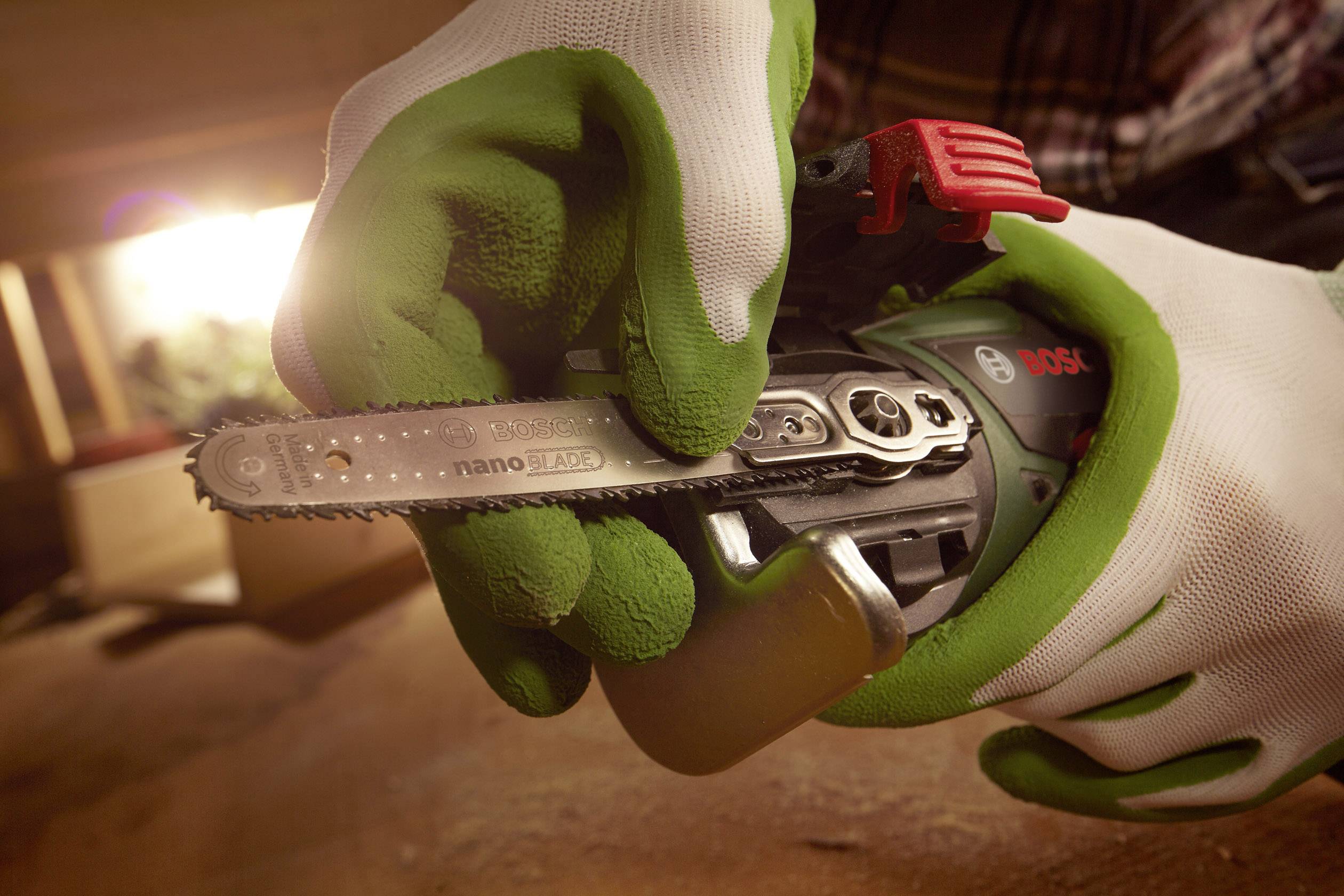 Person mit grünen Handschuhen hält eine kleine Motorsäge, fokussiert auf das Sägeblatt. Umgebung deutet auf handwerkliche Arbeit hin.