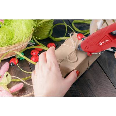 Eine Person verwendet eine Heißklebepistole, um ein Geschenk zu dekorieren. Es gibt Frühlingsdekorationen und bunte Bänder drumherum.