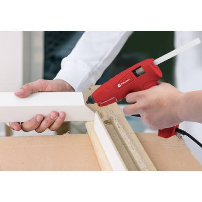 Person verwendet eine rote Heißklebepistole, um ein weißes Stück Holz zu verkleben. Hände stabilisieren das Werkstück auf einer Holzfläche.