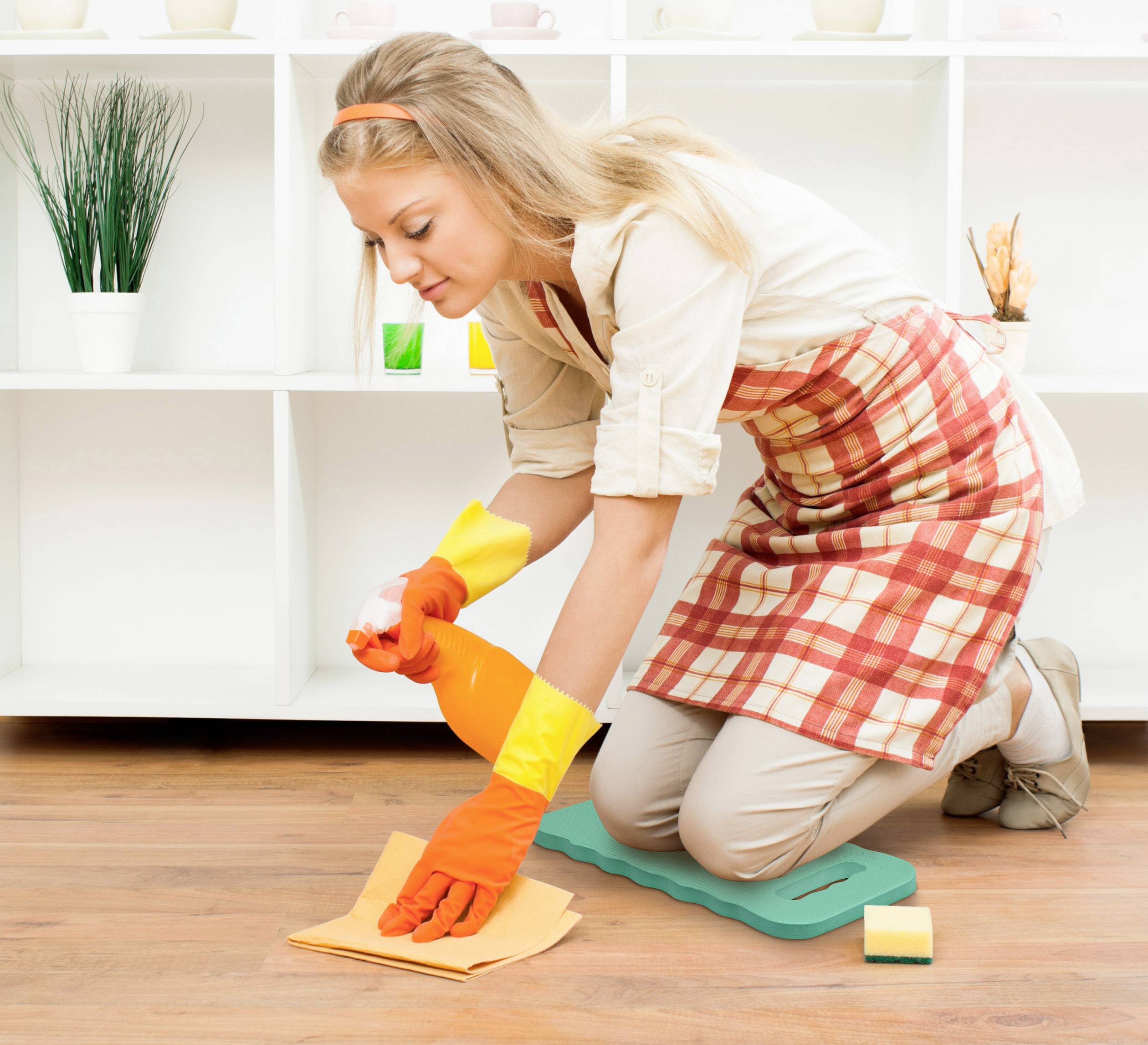 Eine Frau trägt eine gemusterte Schürze und reinigt den Boden mit einem Tuch. Im Hintergrund sind weiße Regale mit Dekor.