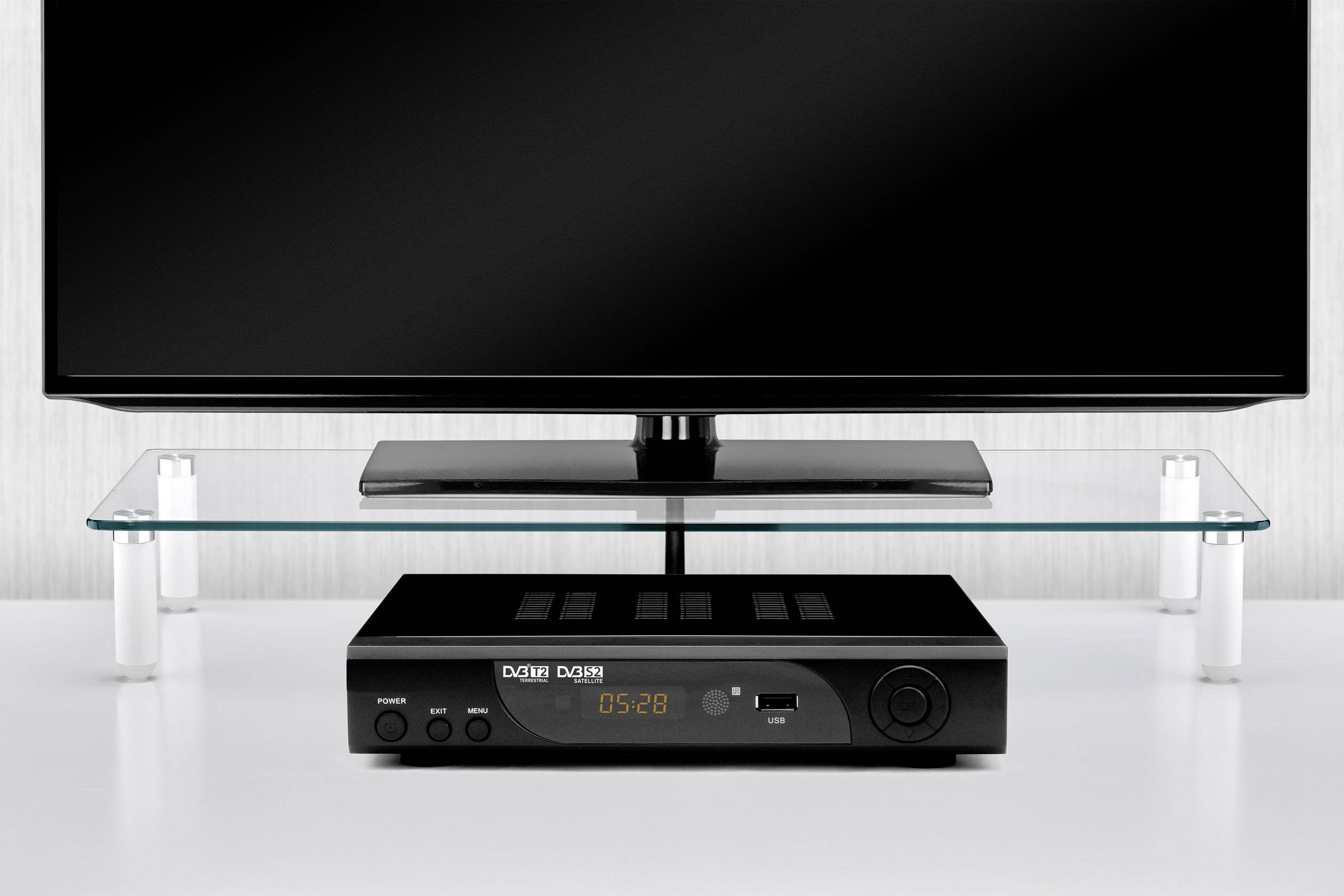 Digitaler Fernsehempfänger vor einem Flachbildschirm, auf einem Glastisch stehend. Gerät zeigt Uhrzeit und Logos an.