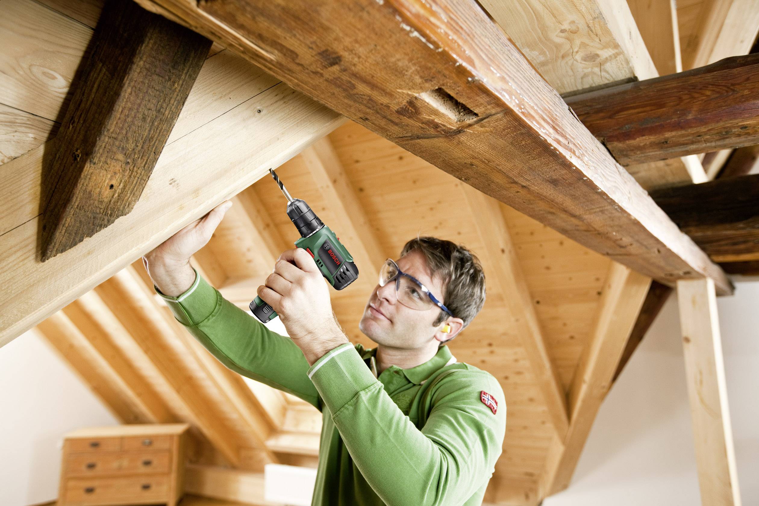Ein Mann in Schutzbrille arbeitet mit einem Akkubohrer an einem Holzbalken in einem Dachstuhl. Er trägt ein grünes Shirt.