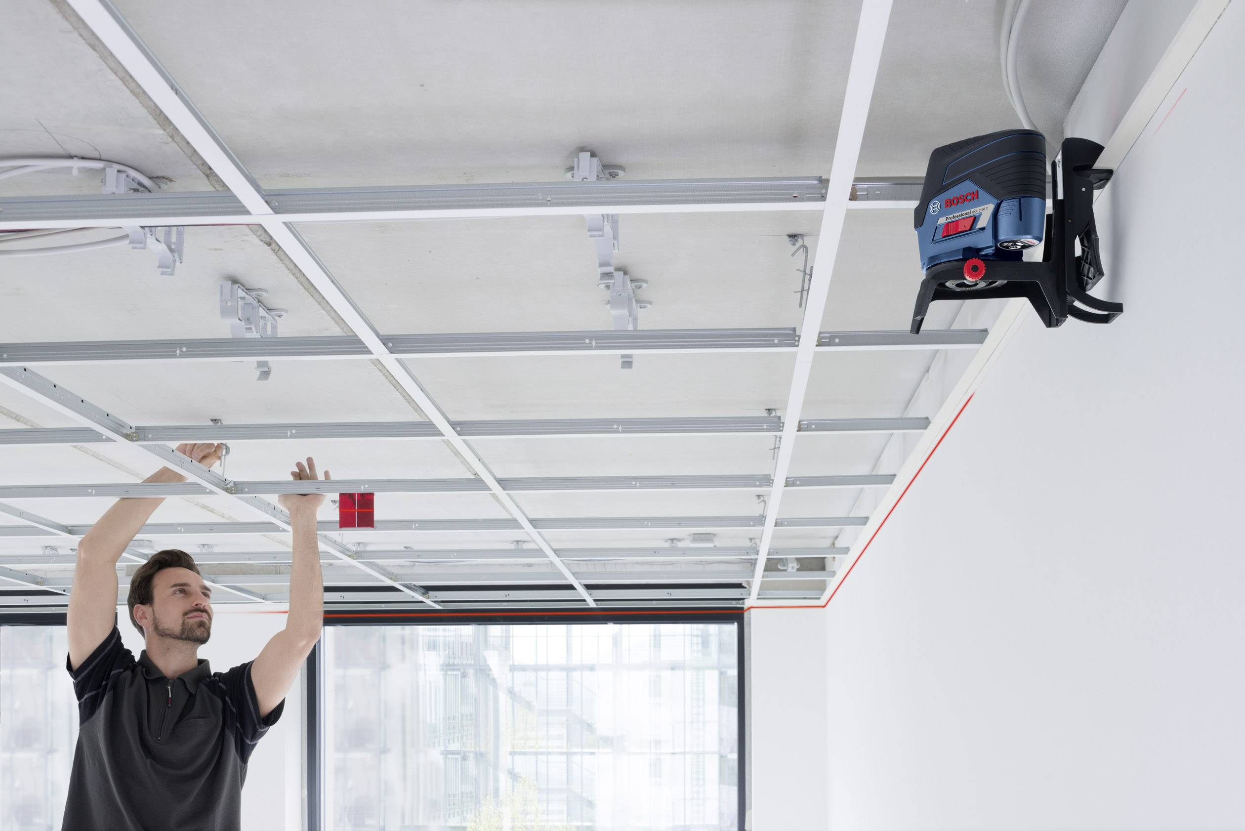 Ein Mann baut eine abgehängte Decke ein und verwendet ein Laser-Nivelliergerät. Er hält eine Messlatte, um die Höhe zu überprüfen.