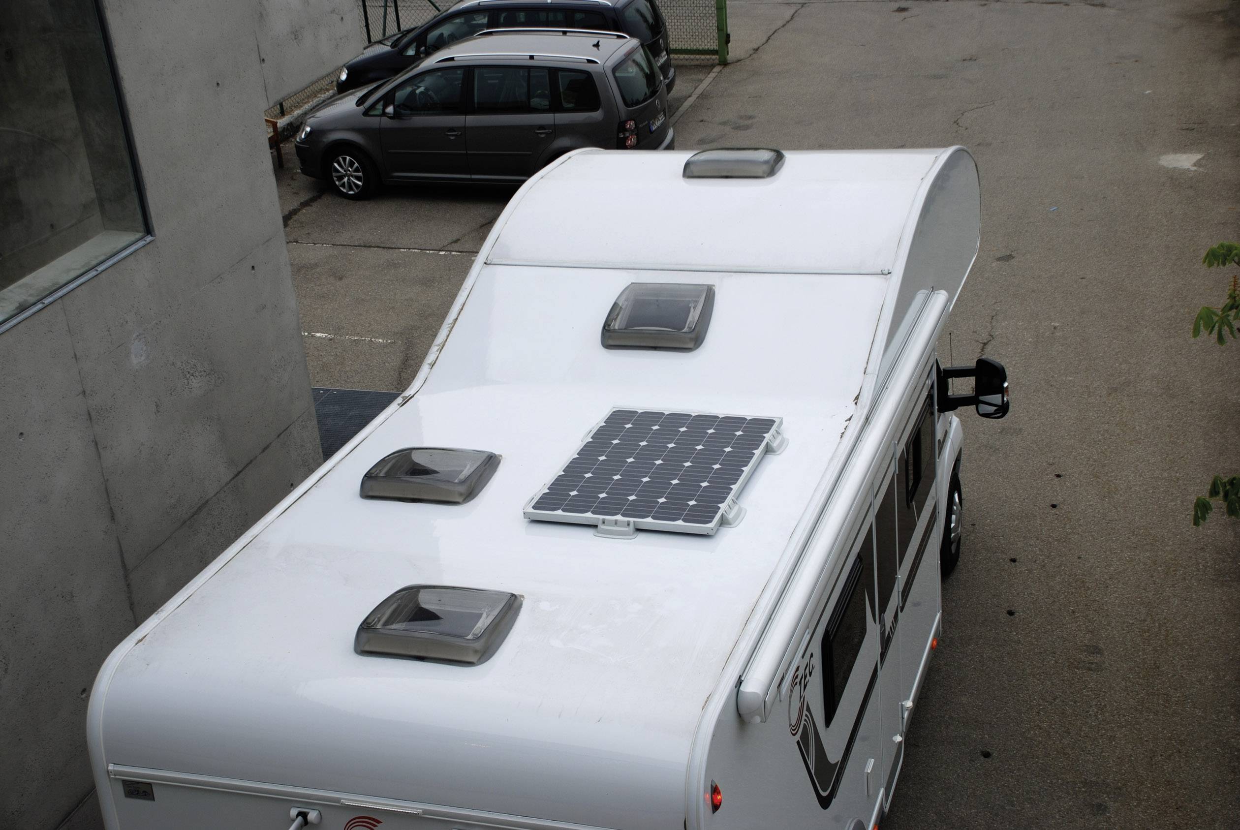 Weißes Wohnmobil mit Solarpanel auf dem Dach, geparkt in einer asphaltierten Einfahrt neben einem Gebäude.