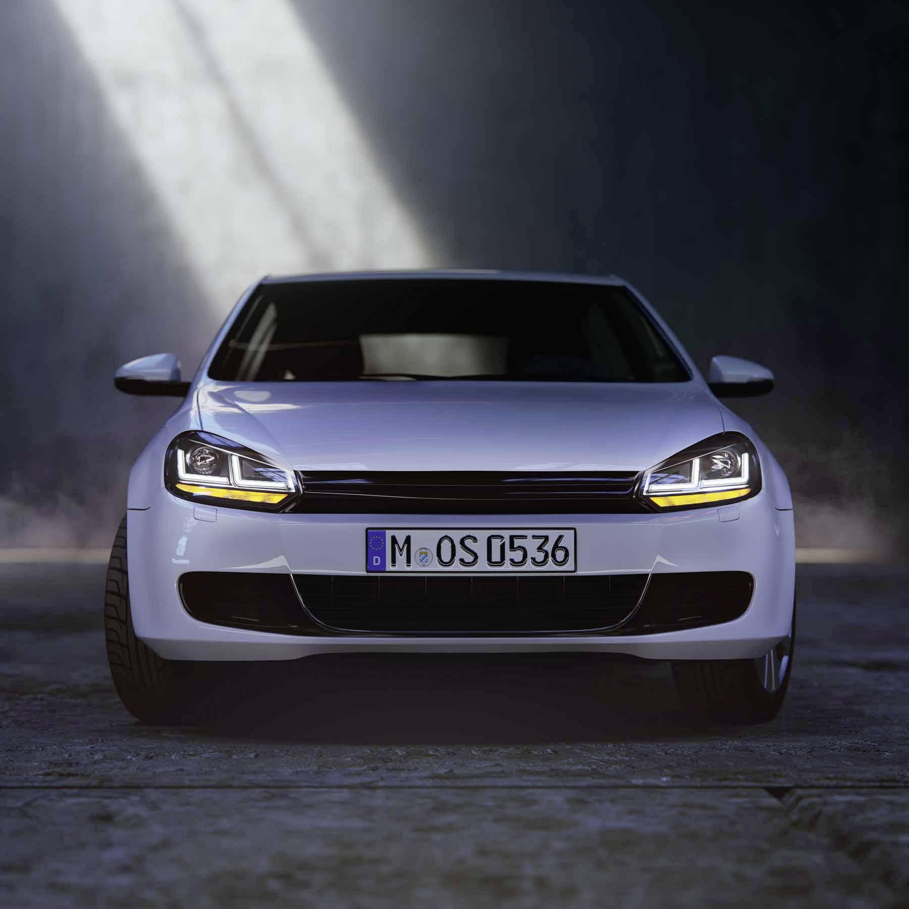 Ein weißes Auto mit eingeschalteten Scheinwerfern steht in einem dunklen Raum. Licht fällt von oben auf das Fahrzeug.