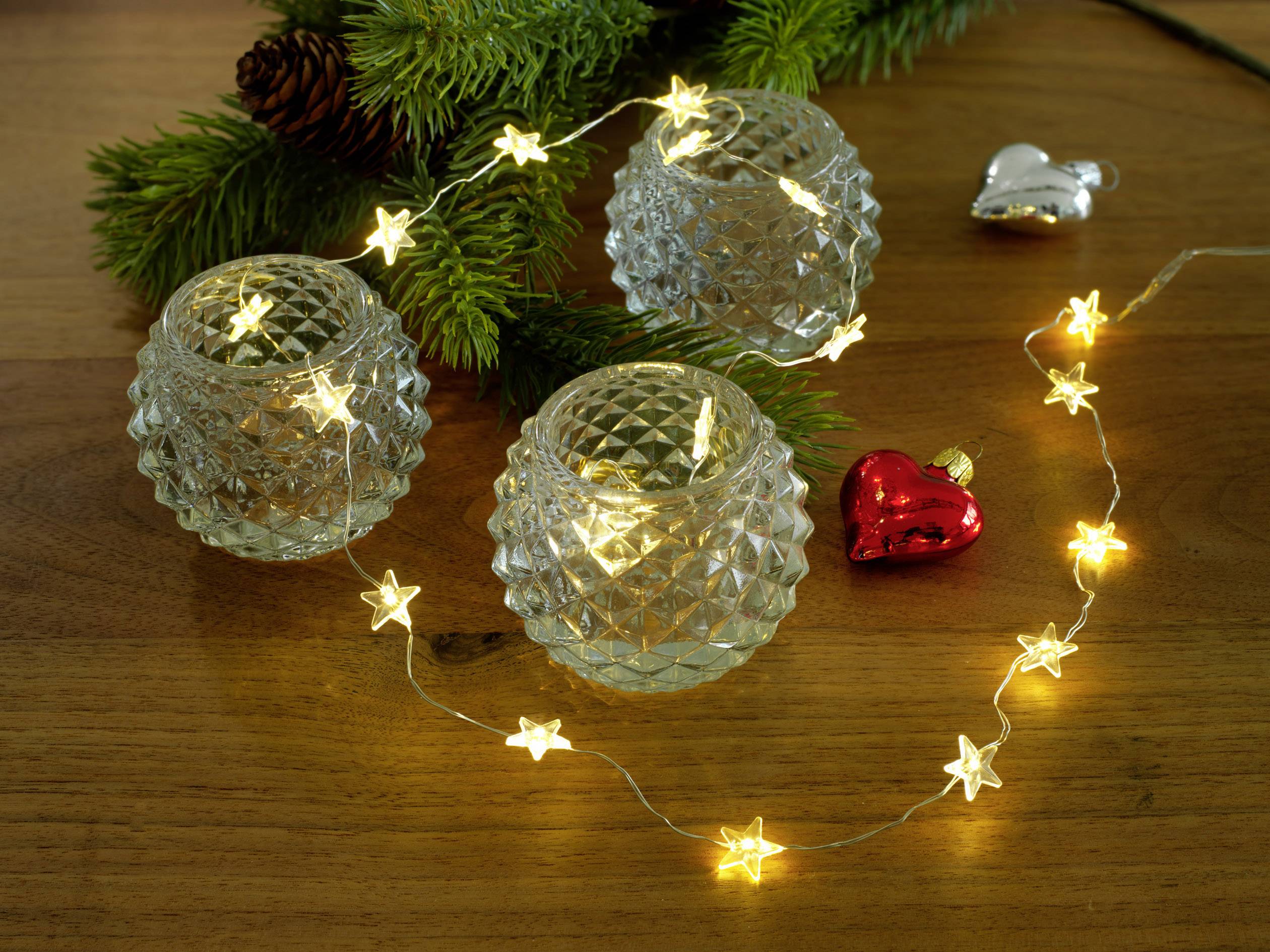 Drei runde, strukturierte Teelichthalter und eine Lichterkette mit Sternen auf einem Holztisch, umgeben von Tannenzweigen und Weihnachtsschmuck.