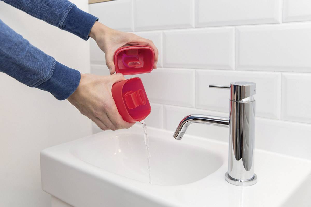 'Person leert roten Plastikbehälter mit Wasser über einem weißen Waschbecken aus. Wasser fließt in das Becken.'