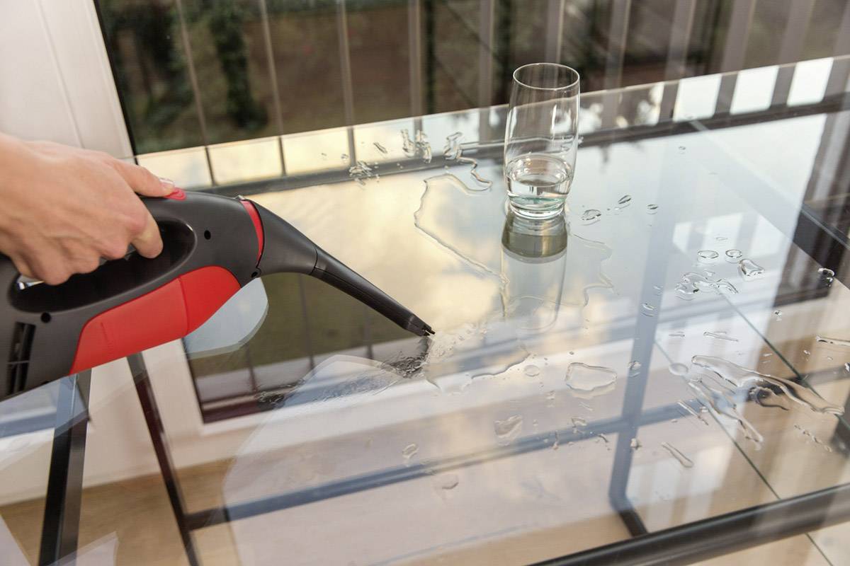 Eine Person benutzt einen Handstaubsauger, um verschüttetes Wasser auf einem Glastisch neben einem halb vollen Glas zu reinigen.
