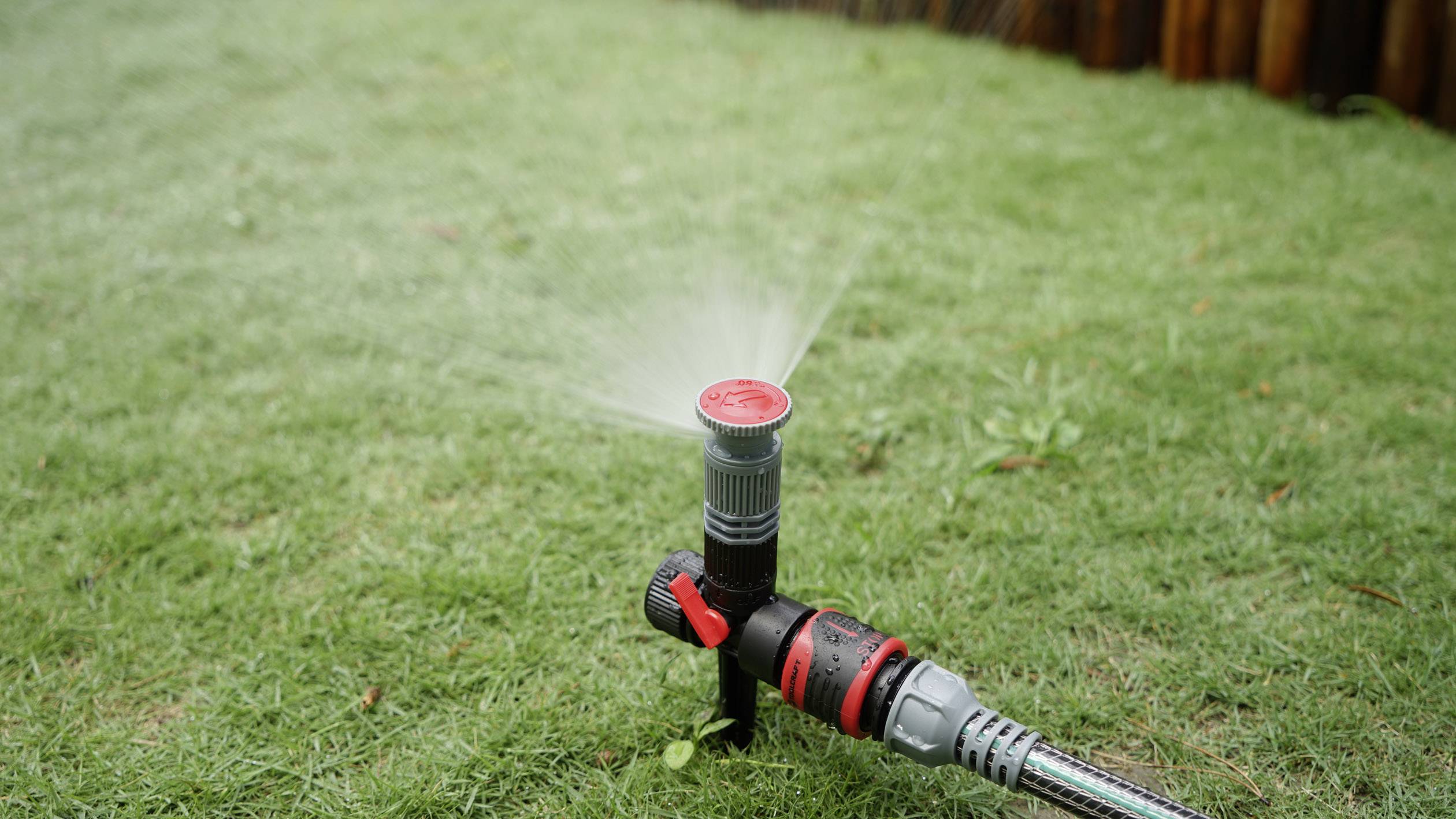 Ein Sprinkler auf einer Rasenfläche verteilt gleichmäßig Wasser. Das Gras ist saftig grün, und es gibt Holz im Hintergrund.