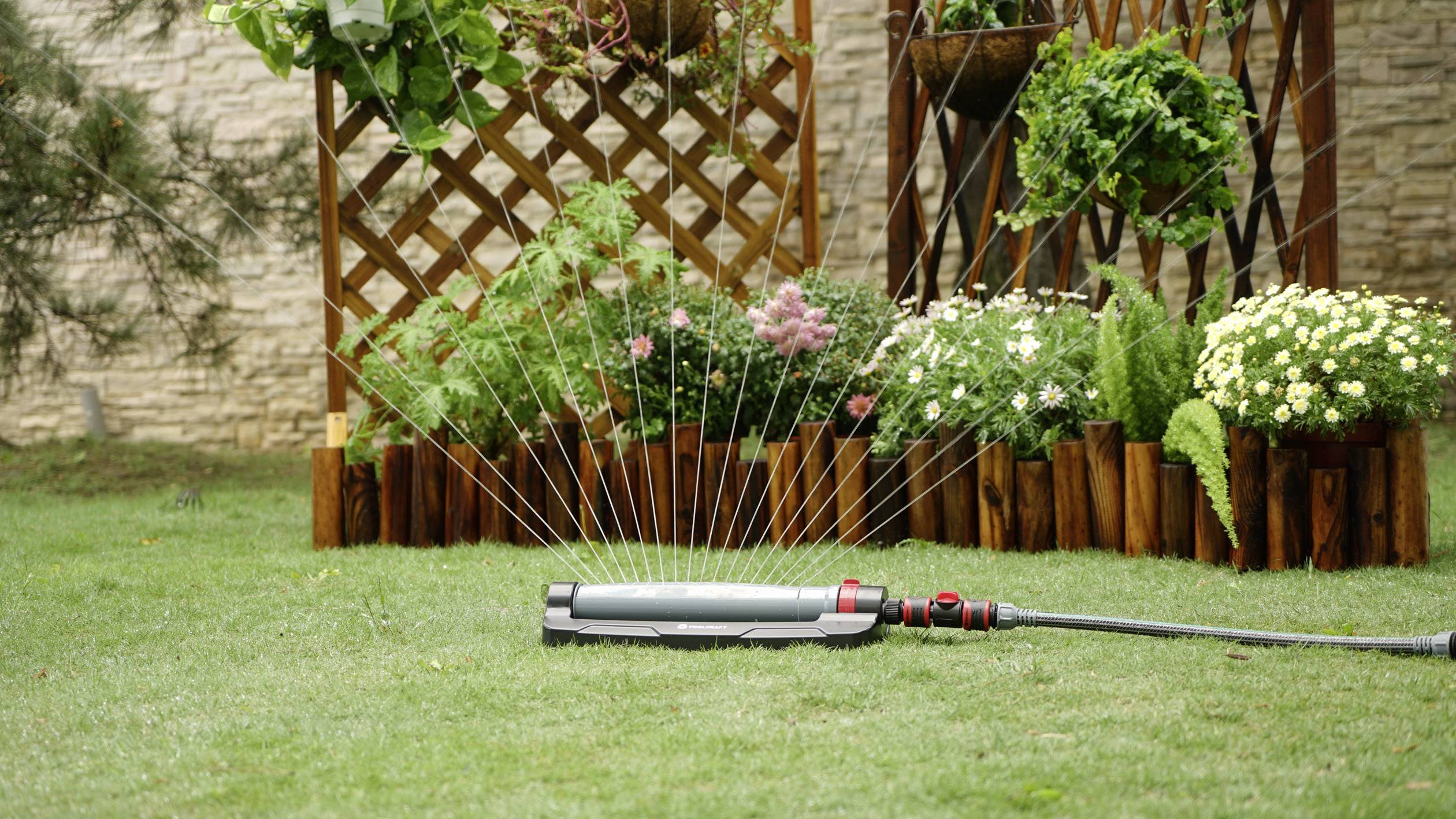 Ein Rasensprinkler bewässert einen grünen Rasen vor einem Holzgitter mit hängenden Pflanzen und Blumenkästen voller blühender Blumen.