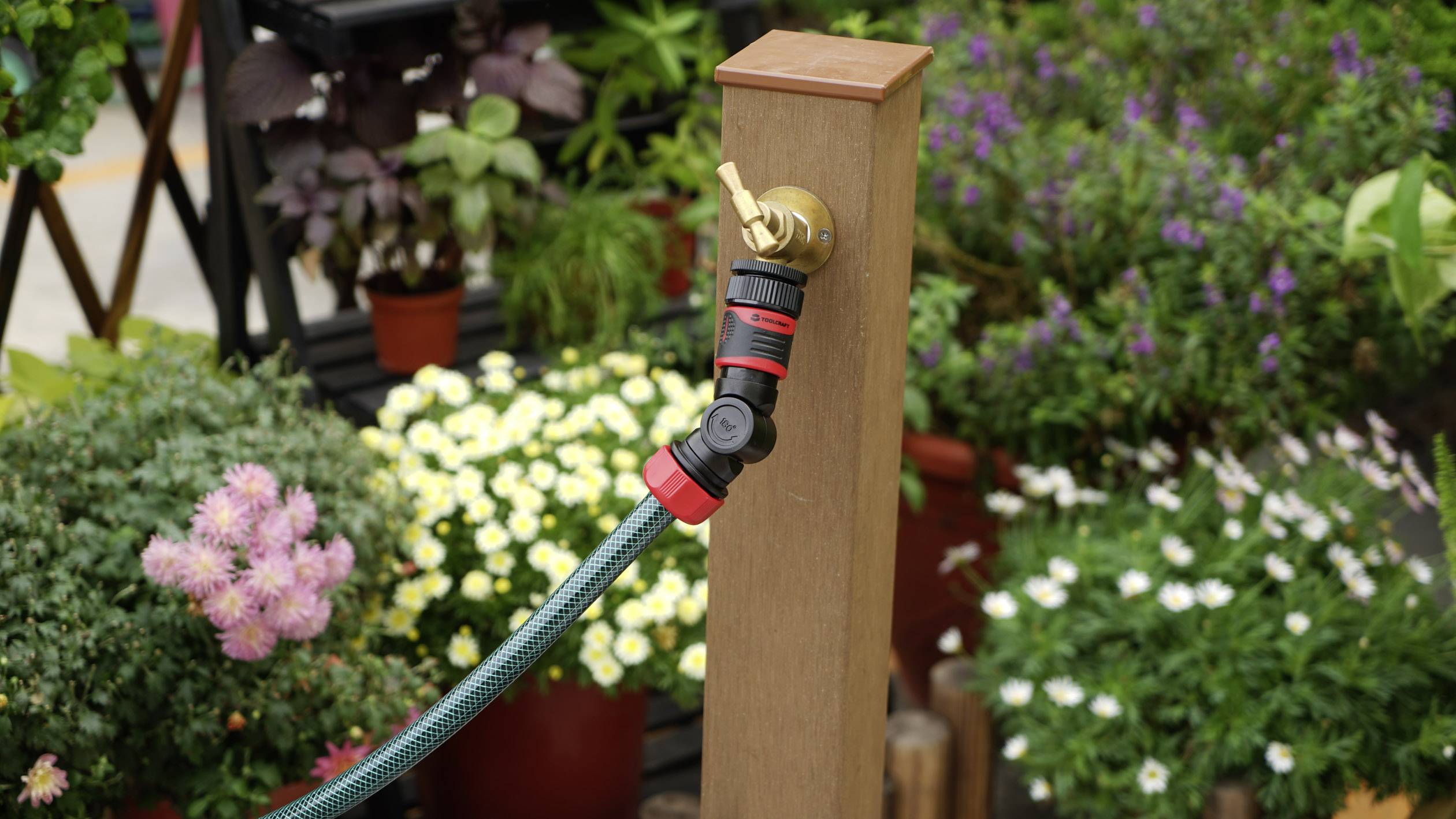 'Gartenschlauch mit Messinganschluss an Holzpfosten befestigt; im Hintergrund blühende Pflanzen.'