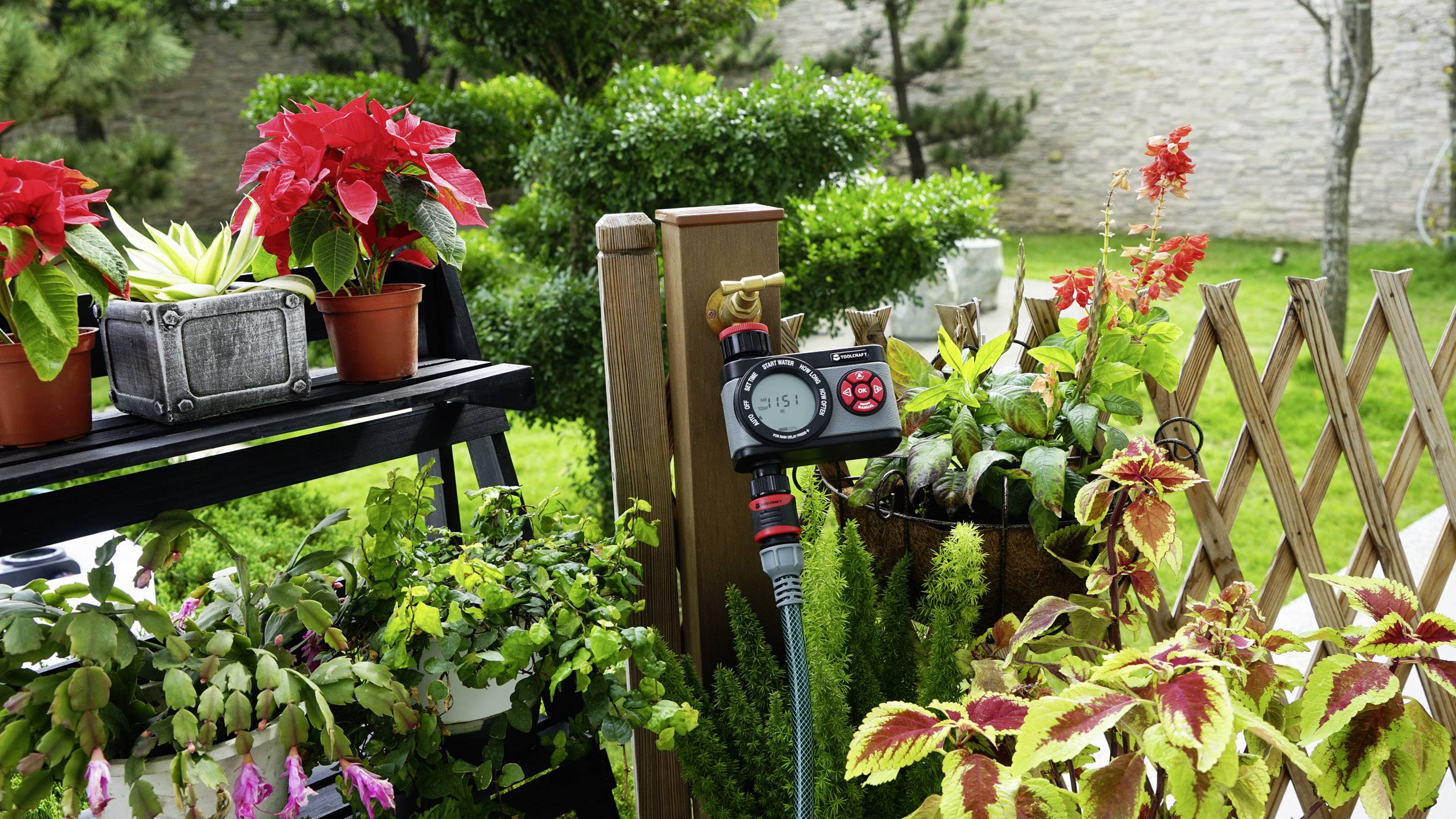 Ein bunter Gartenbereich mit verschiedenen Topfpflanzen und einem Bewässerungssystem-Controller in der Mitte, umgeben von grünem Rasen.