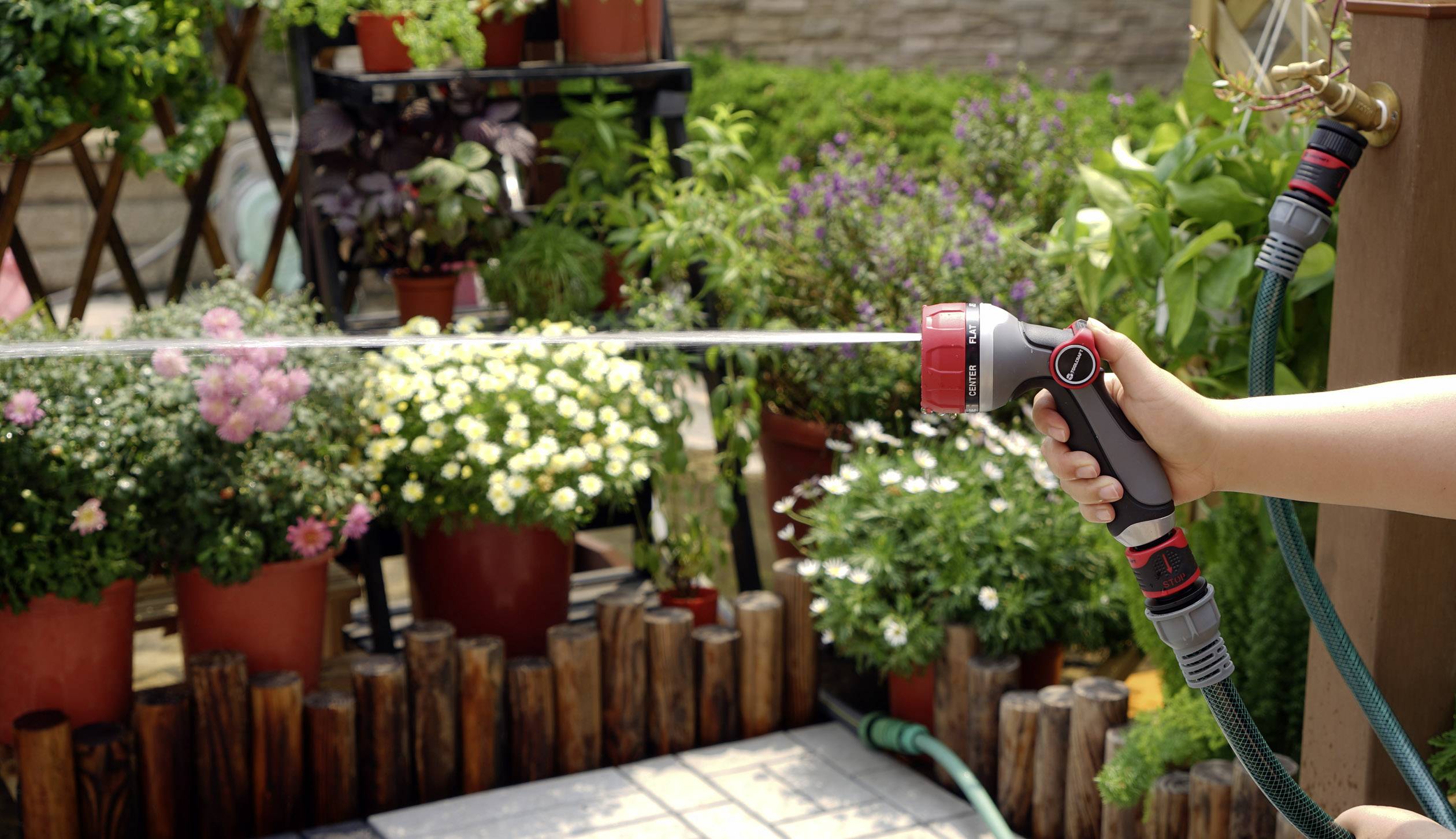 Eine Person gießt Pflanzen im Garten mit einem Wasserschlauch. Im Hintergrund sind viele grüne und blühende Topfpflanzen zu sehen.