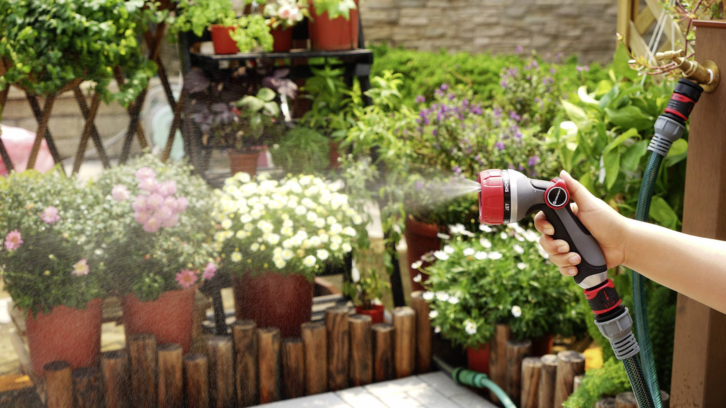 Eine Person gießt einen blühenden Garten mit einem Gartenschlauch. Verschiedene Blumen in Töpfen stehen auf einer Holzterrasse.