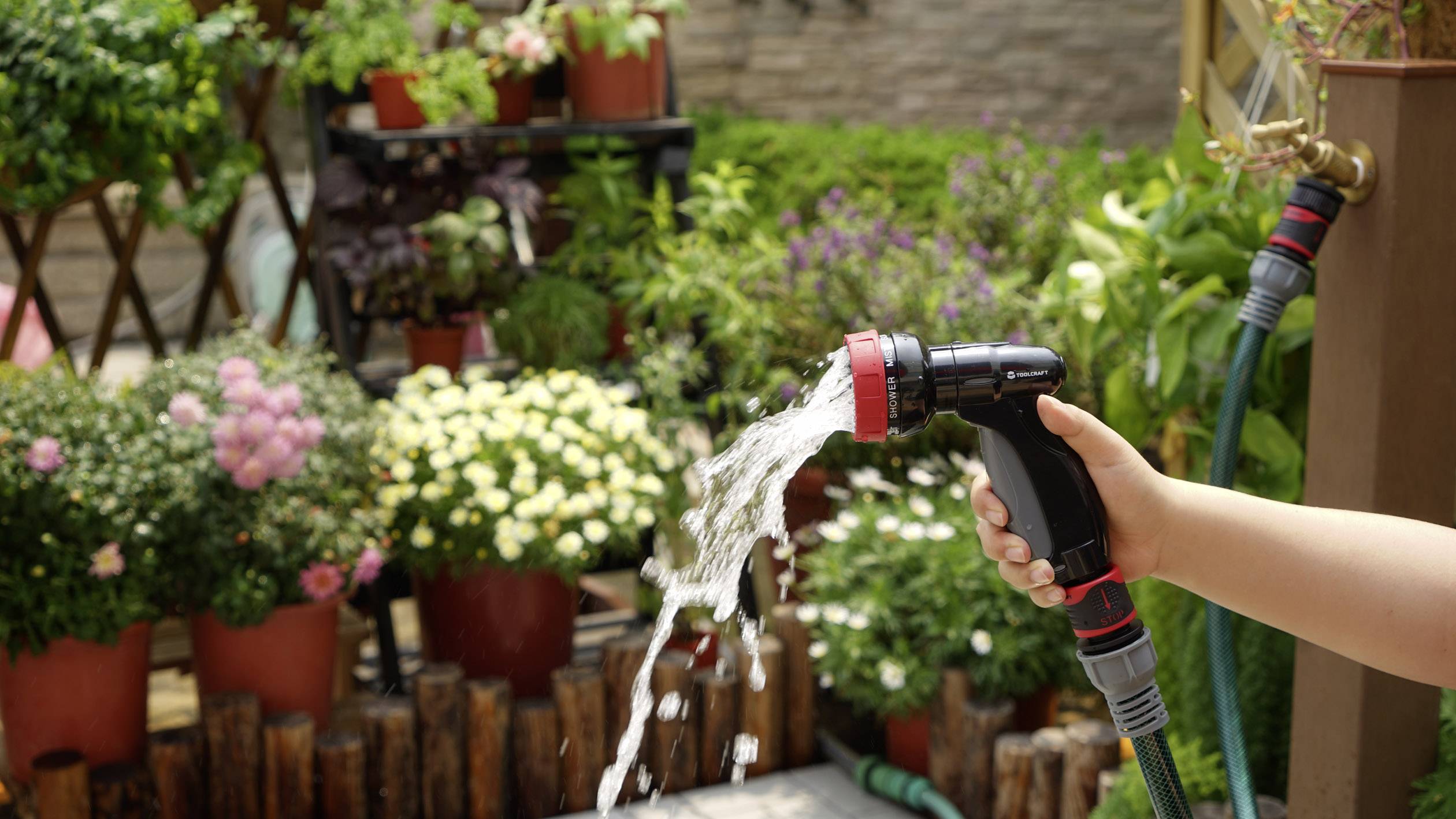 Eine Person gießt mit einem Wasserschlauch eine Vielzahl blühender Pflanzen in Töpfen in einem üppigen Garten.
