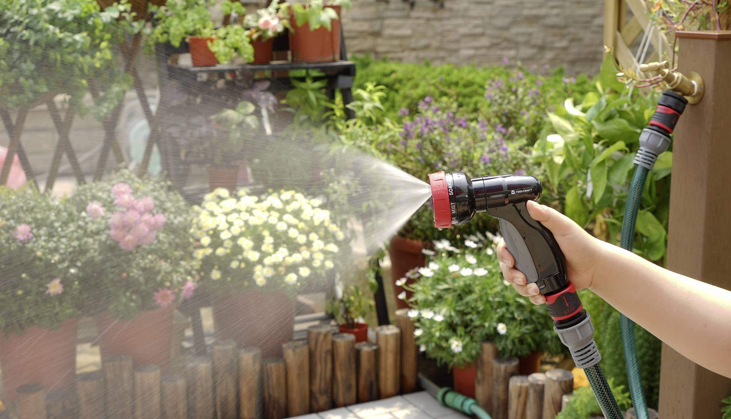 Eine Person gießt mit einem Gartenschlauch Pflanzen in einem blühenden Garten. Im Hintergrund sind Topfpflanzen zu sehen.