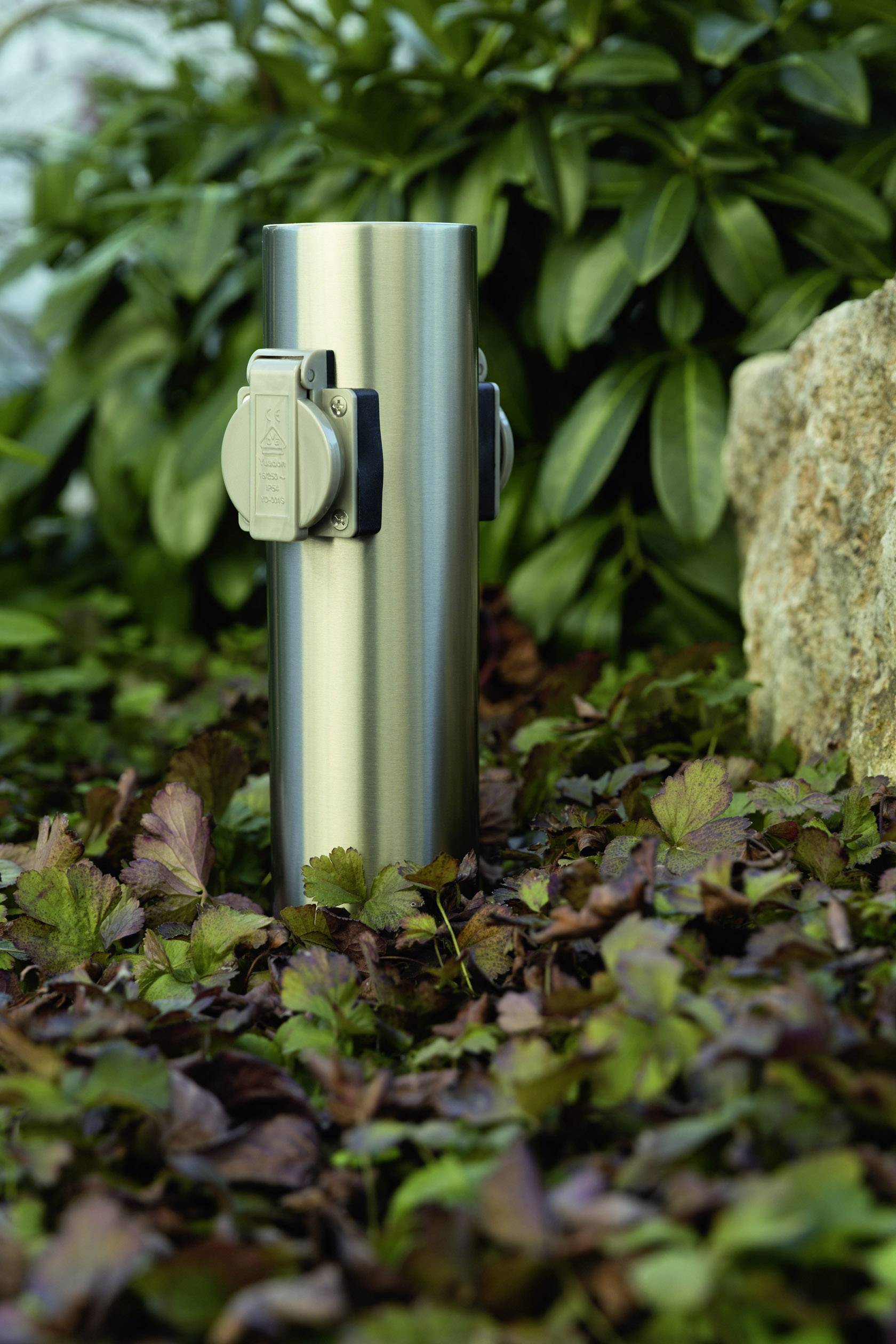 Eine Edelstahl-Gartensteckdose mit zwei Steckdosen steht auf einem Boden mit Blättern, umgeben von grünen Pflanzen und einem Stein.