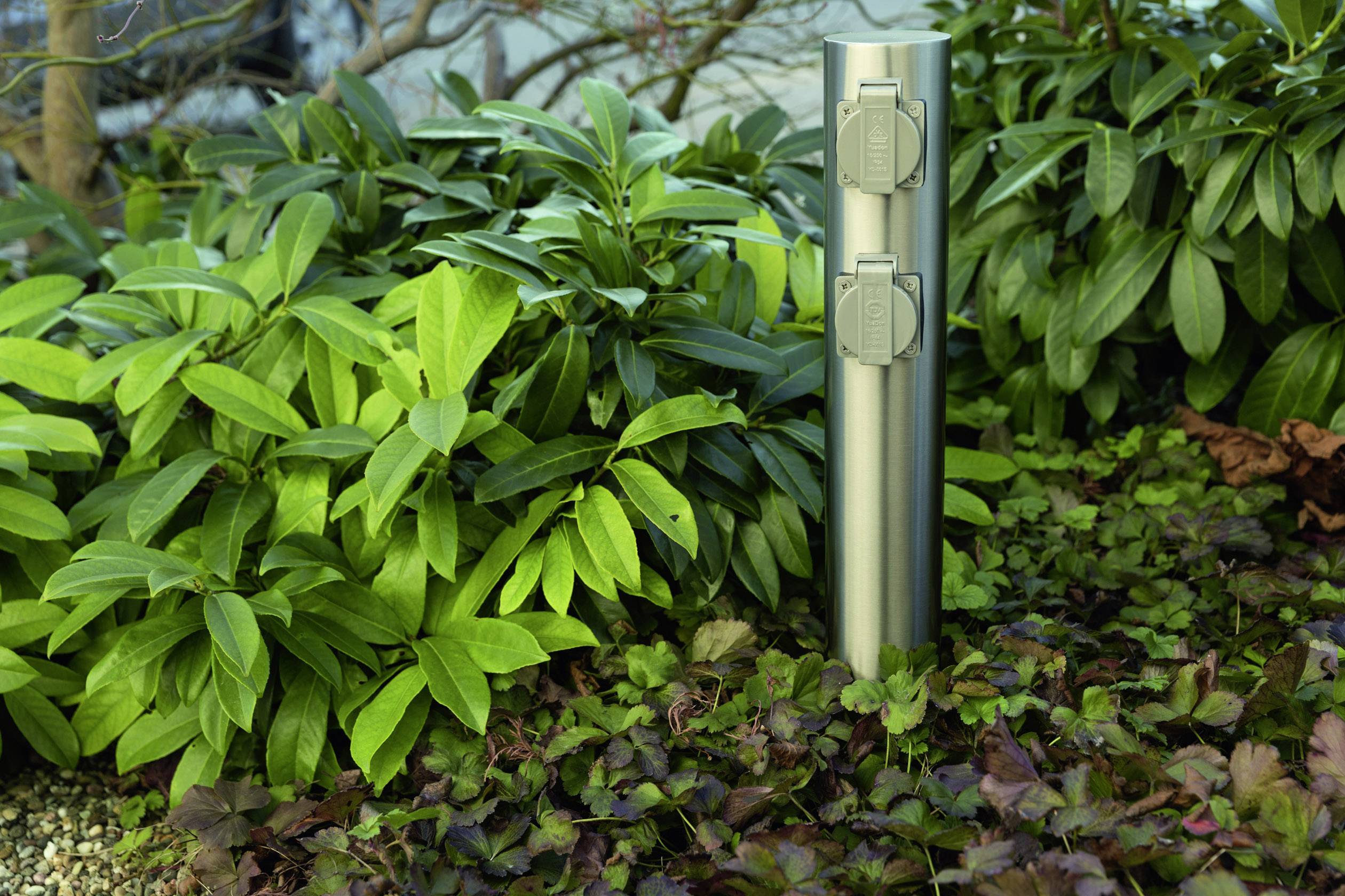 Ein silberner Außensteckdosensockel mit zwei Steckplätzen steht zwischen dichten, grünen Blättern und Pflanzen im Garten.