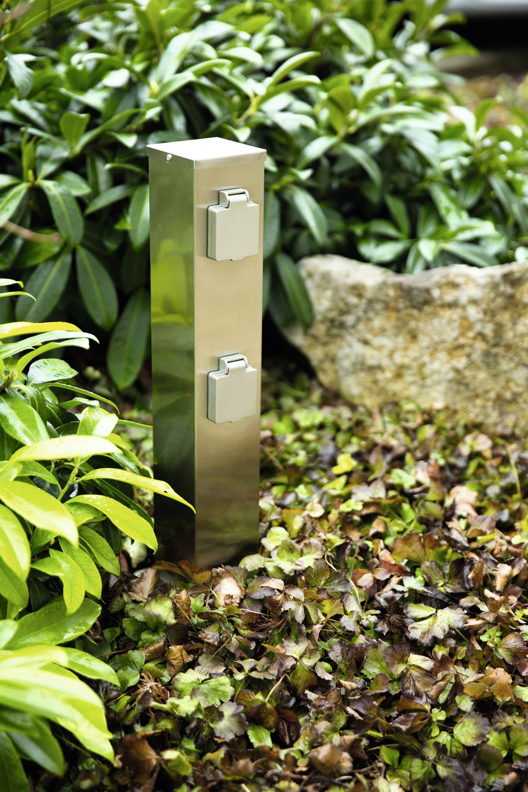 Metallische Outdoor-Steckdosensäule mit zwei Steckdosen in einem Garten, umgeben von Laub und Pflanzen. Ideal für Gartenarbeiten.