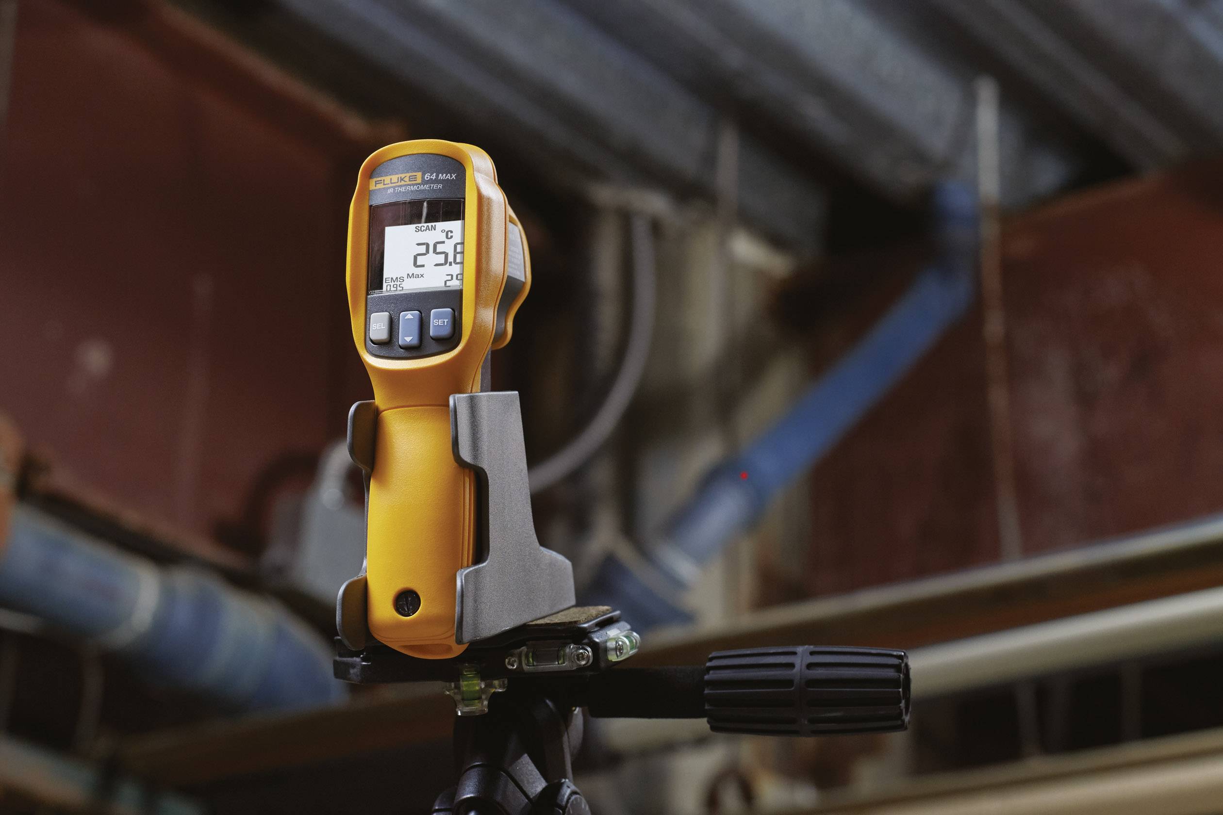Ein gelbes Infrarotthermometer auf einem Stativ zeigt 25,2 Grad an. Hintergrund zeigt industrielle Rohre und Metallkonstruktionen.
